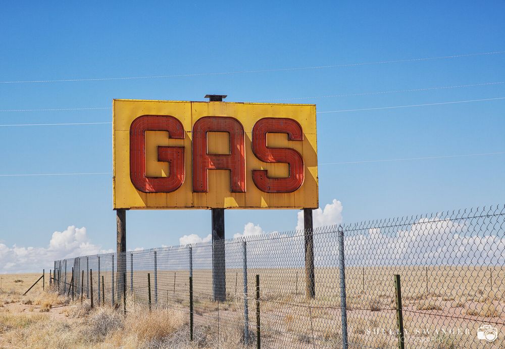 Retro yellow GAS sign stands alone in desert landscape behind chain link fence.