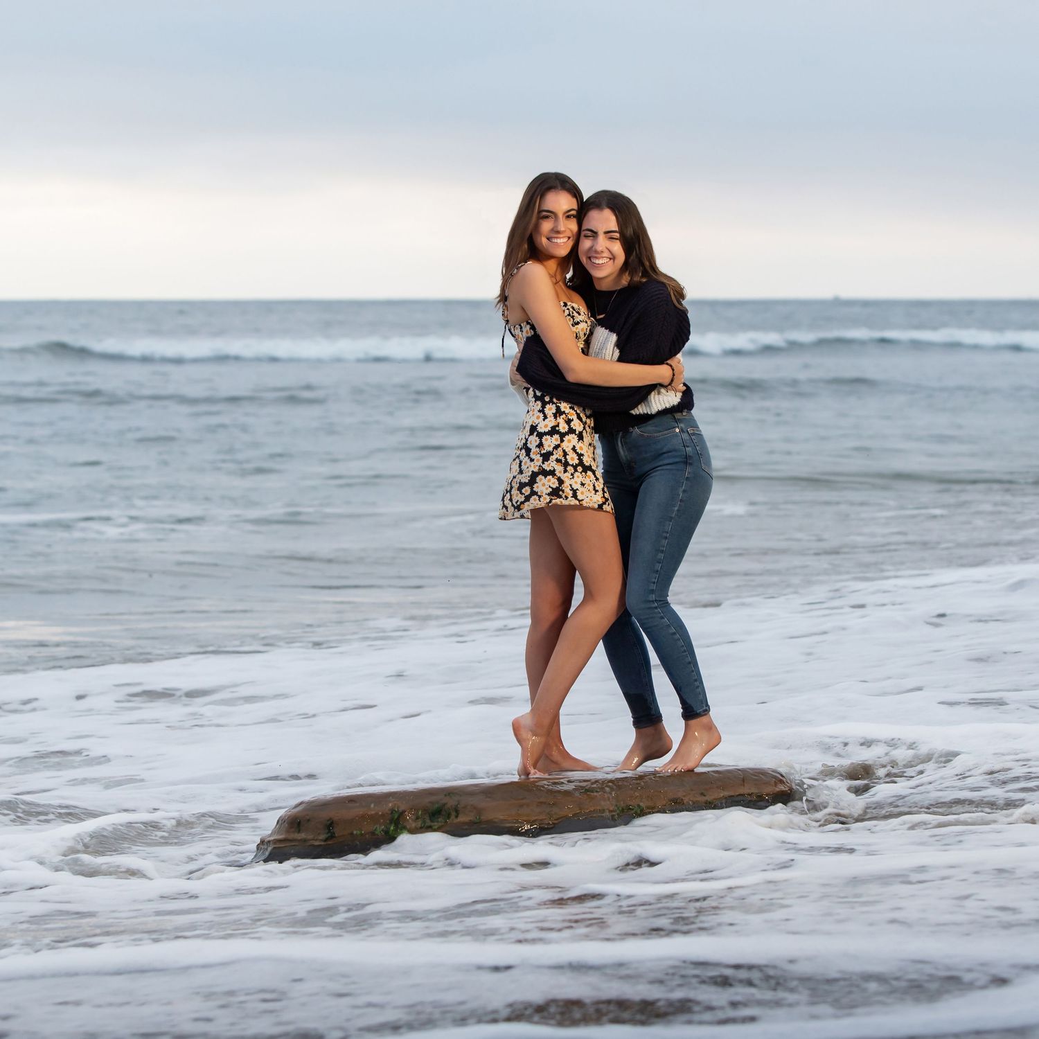 Two people embrace while standing on a rock at the beach with waves in the background.