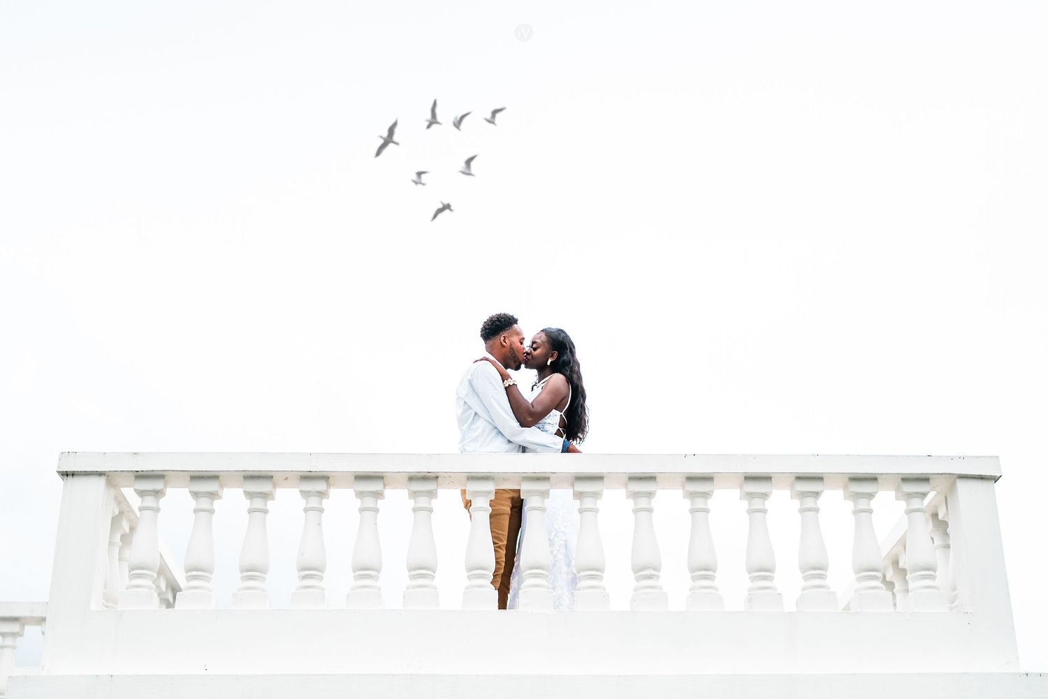 A couple embraces on a white balcony railing with birds flying overhead against a bright sky.