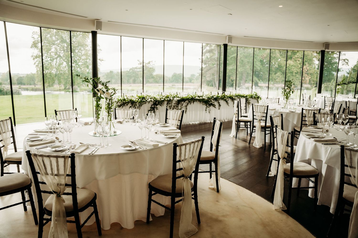 Modern reception venue with floor-to-ceiling windows, white tablecloths, and natural light overlooking green landscape.