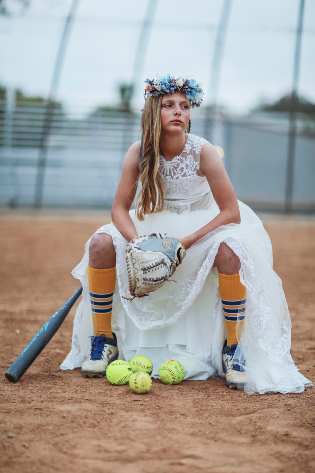 Softball player in white lace wedding dress and yellow socks poses with glove and balls on baseball diamond at sunset.