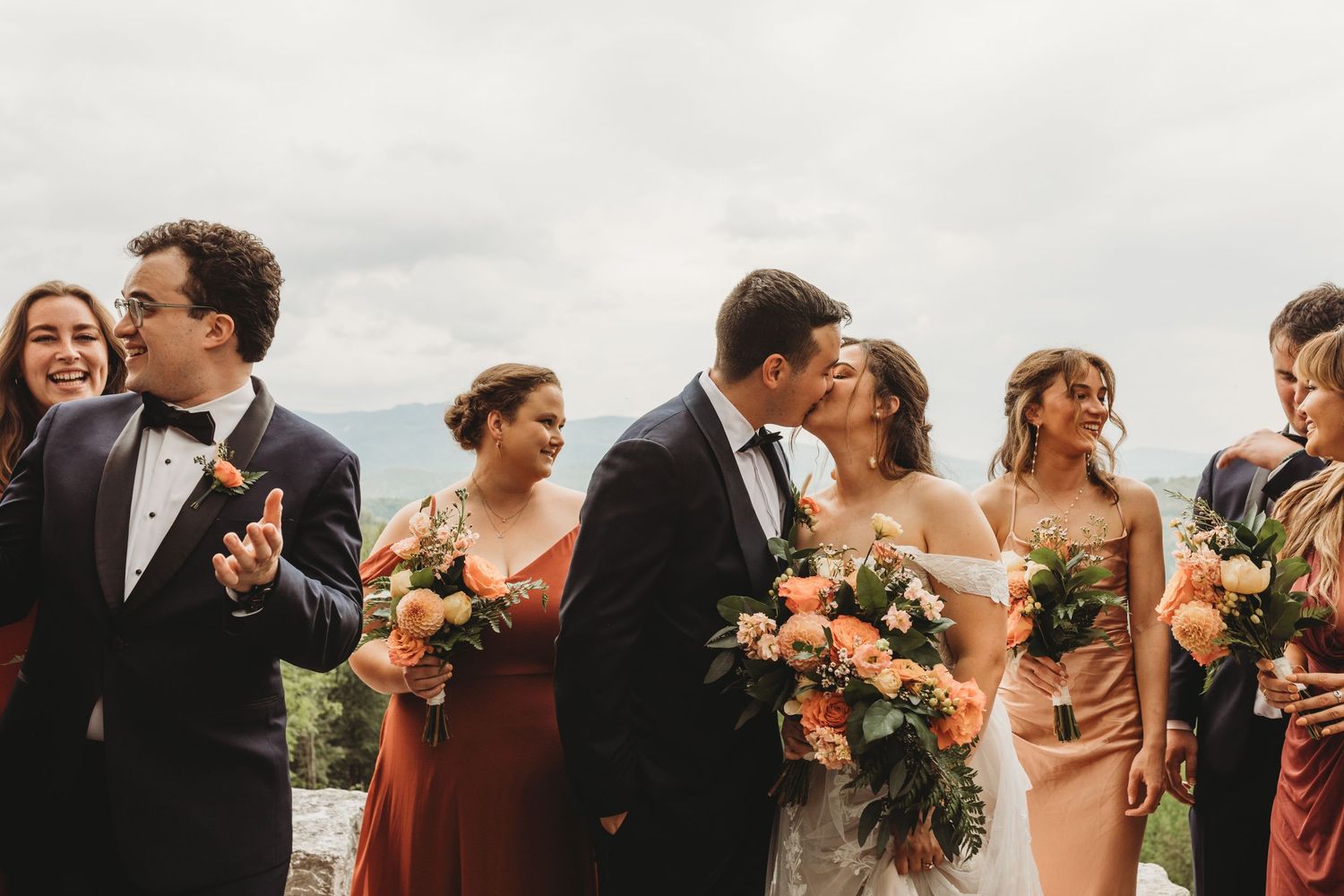 Candid moment of wedding party laughing together outdoors in formal attire with peach floral bouquets.