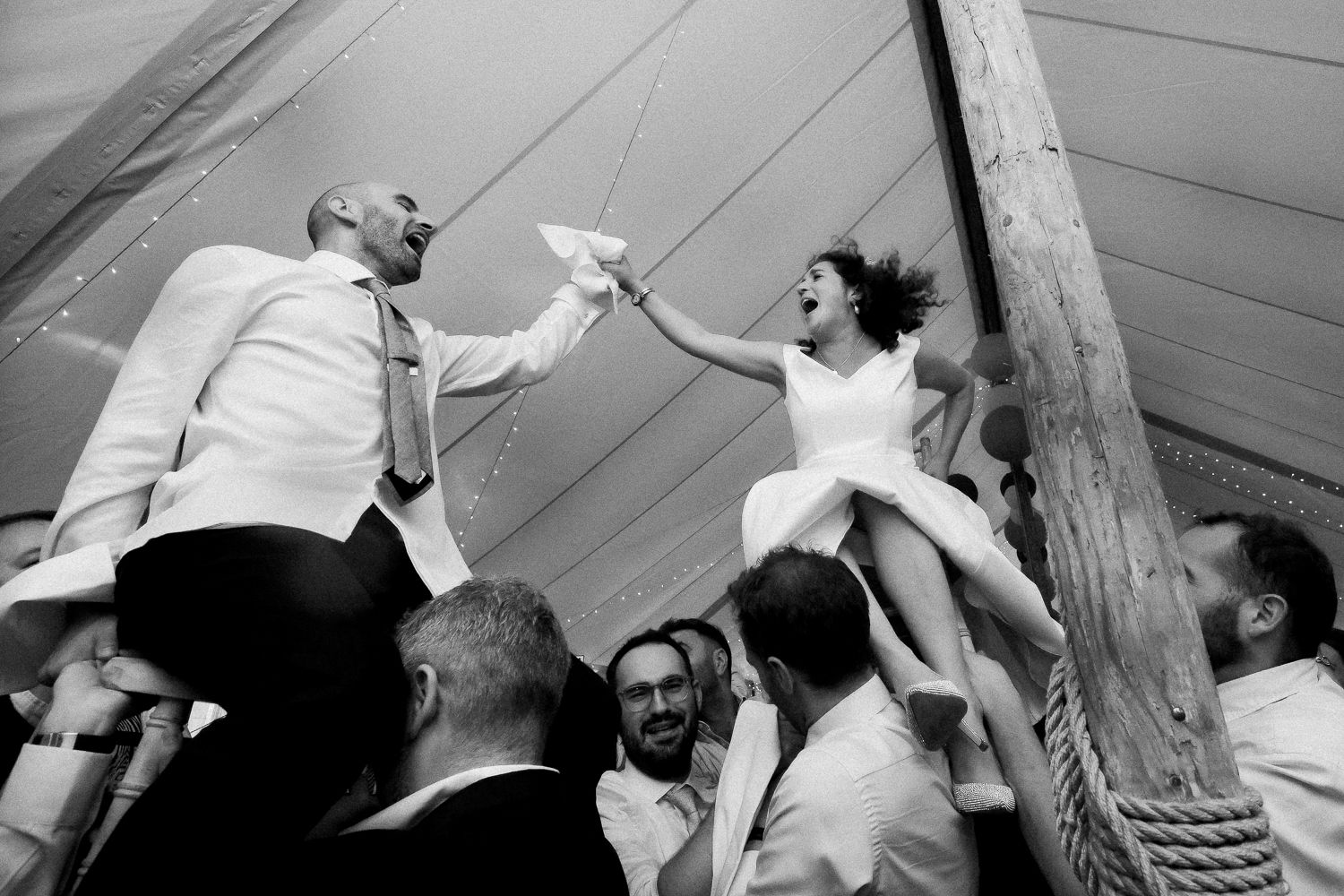 Black and white photo of wedding guests hoisting newlyweds during reception celebrations.