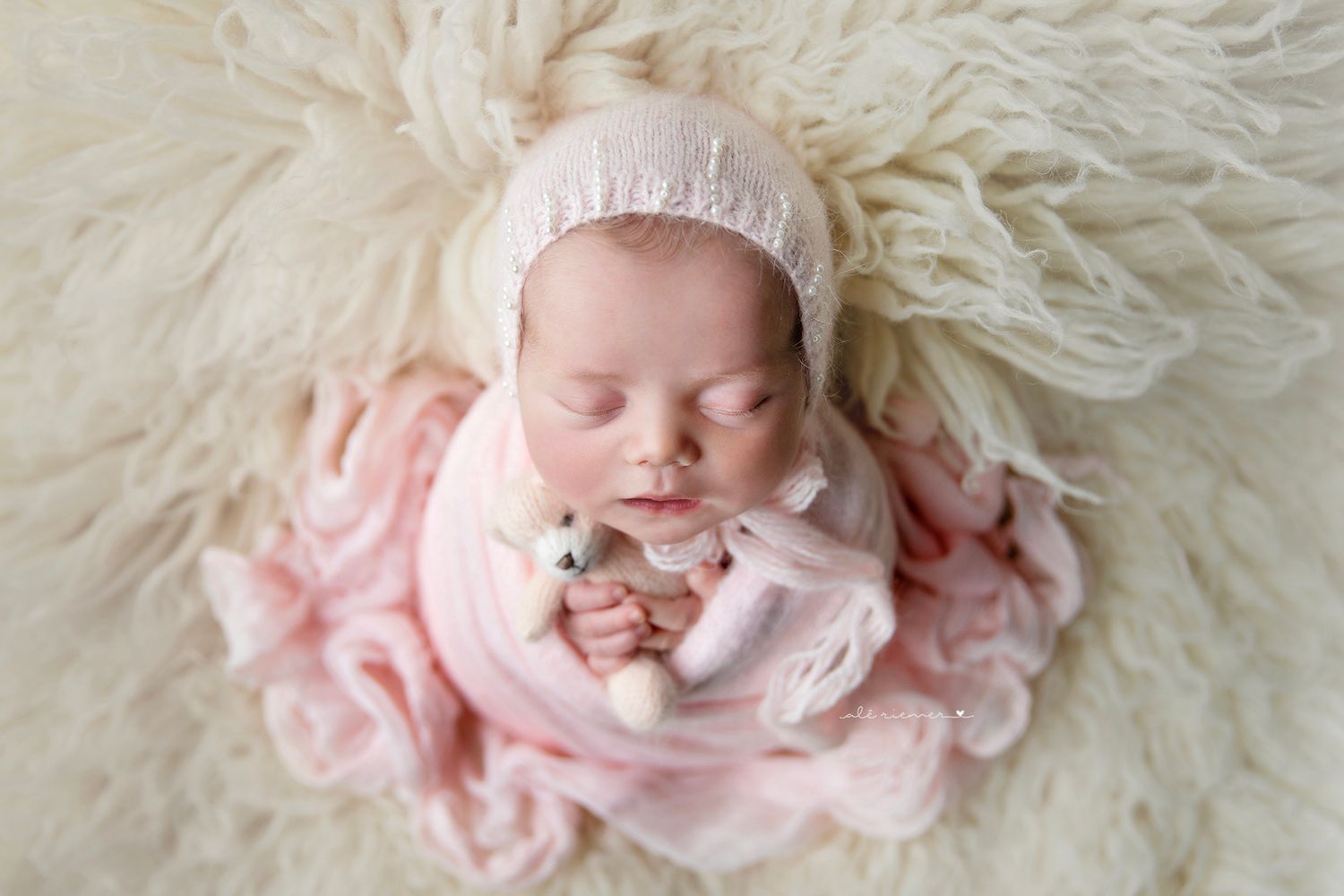 Newborn baby wrapped in soft pink fabric lying on fluffy white blanket wearing delicate bonnet.