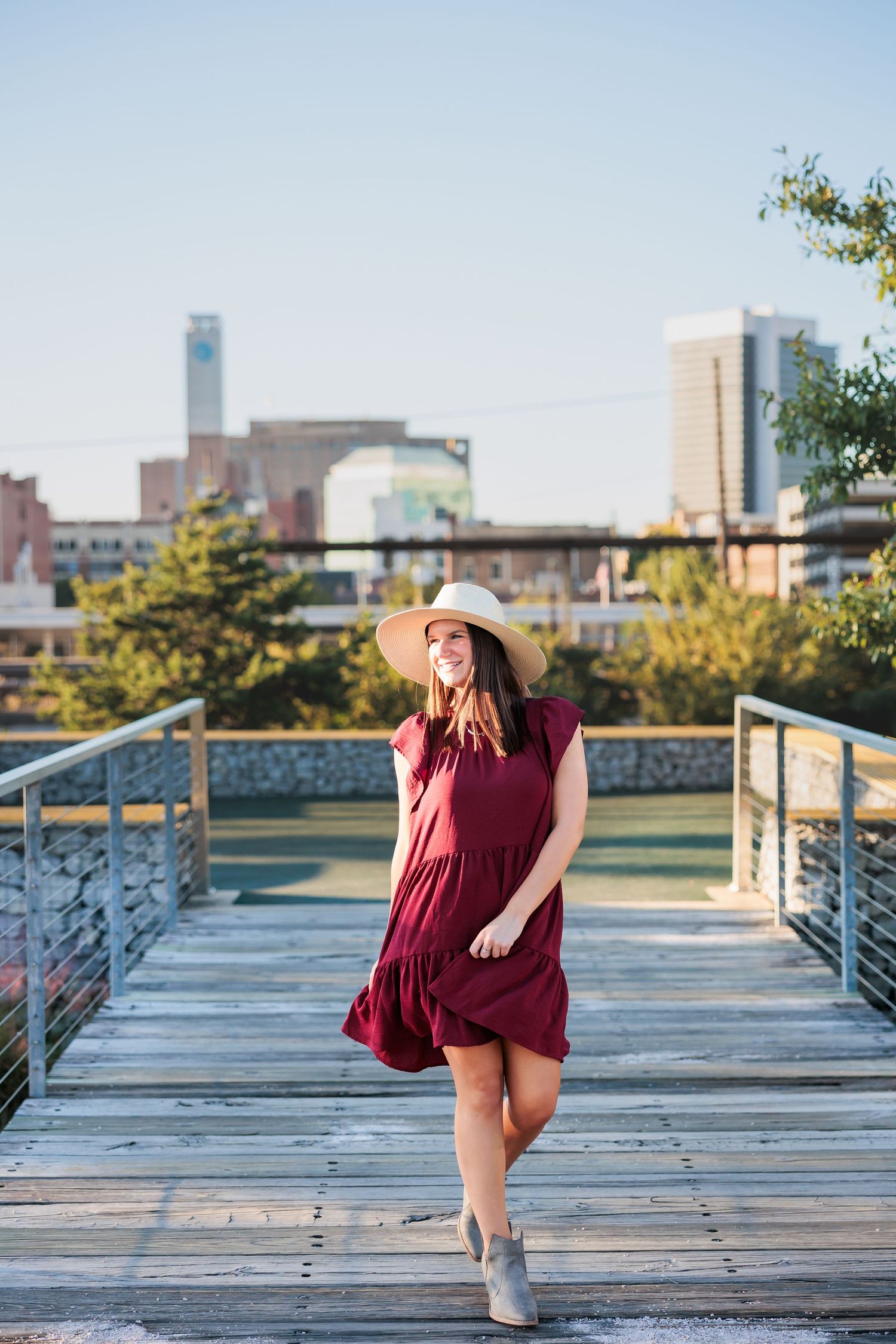 Woman in burgundy dress and hat poses on wooden boardwalk with city skyline behind her.