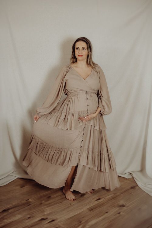A long flowing beige maternity dress with ruffled tiers and button details photographed in a studio setting.