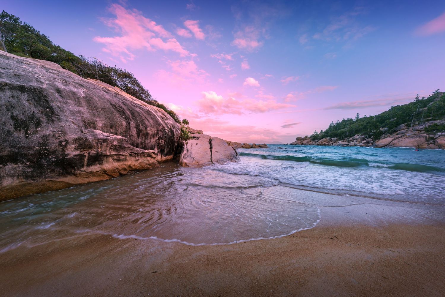 Pink and purple sunrise over rocky beach with waves washing up on shore.