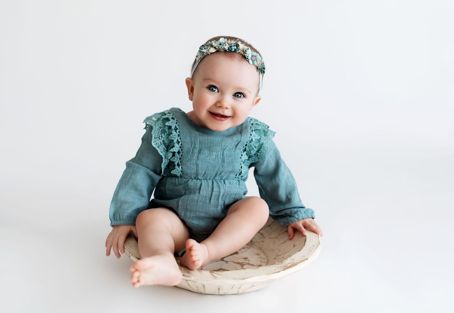 Baby in teal ruffle dress and floral headband sitting on round cushion against white background.