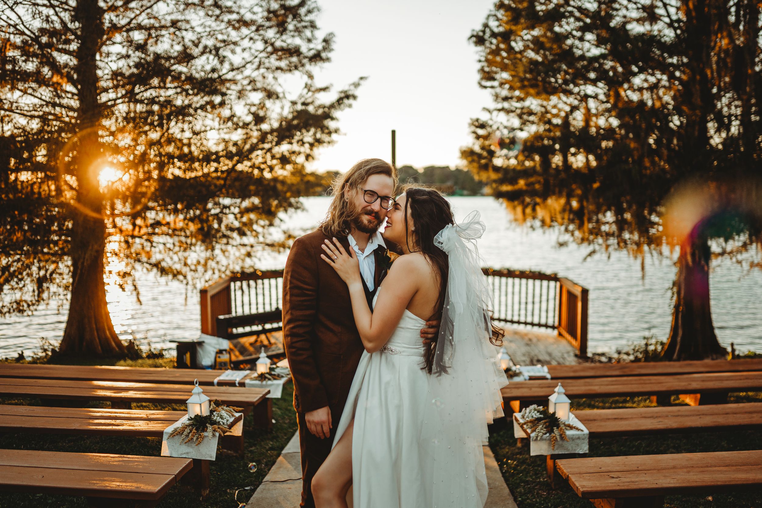 Golden hour wedding portrait by the water with sunburst through trees.
