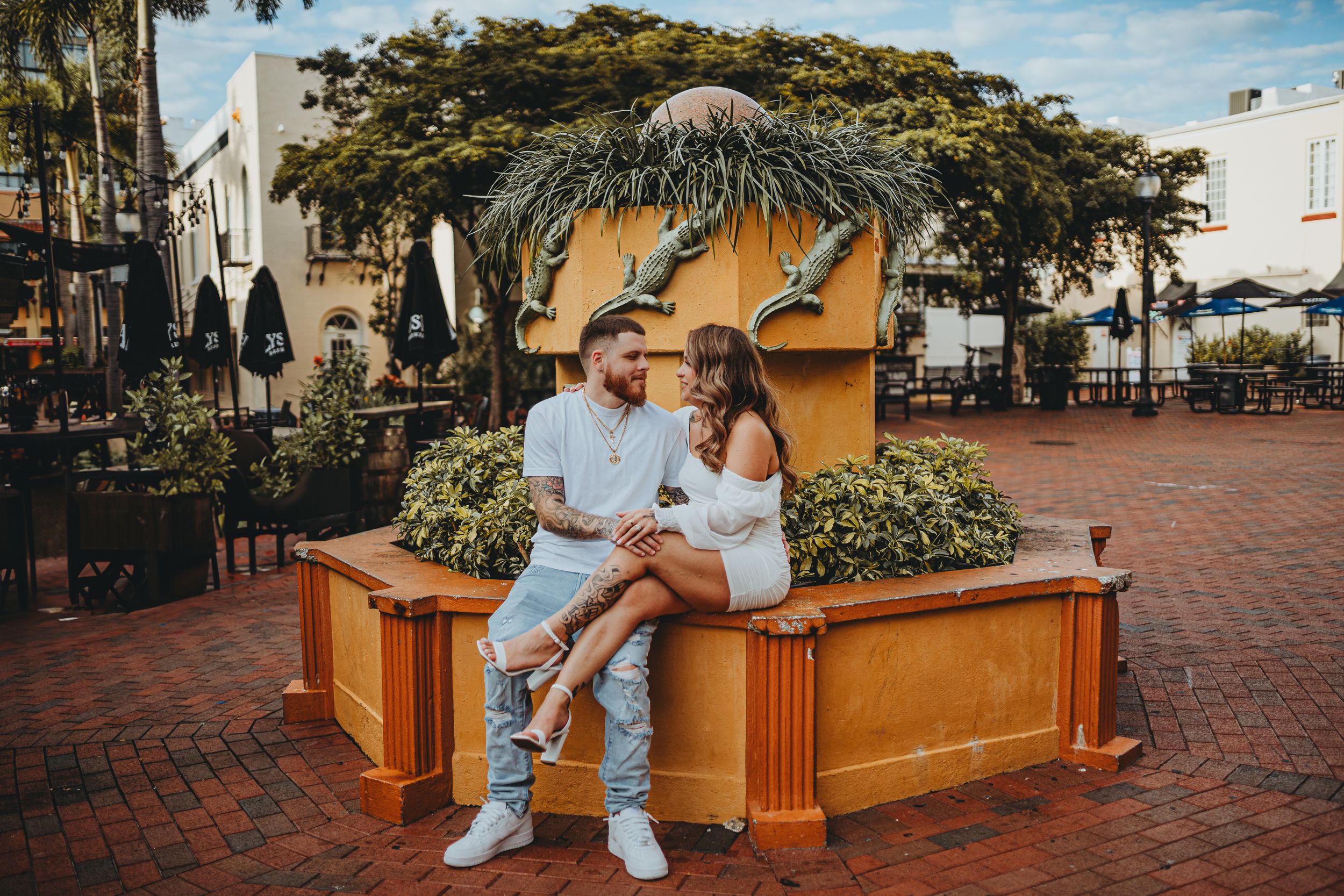 A romantic pair sits on a brick planter with palm trees and vintage architecture in the background.