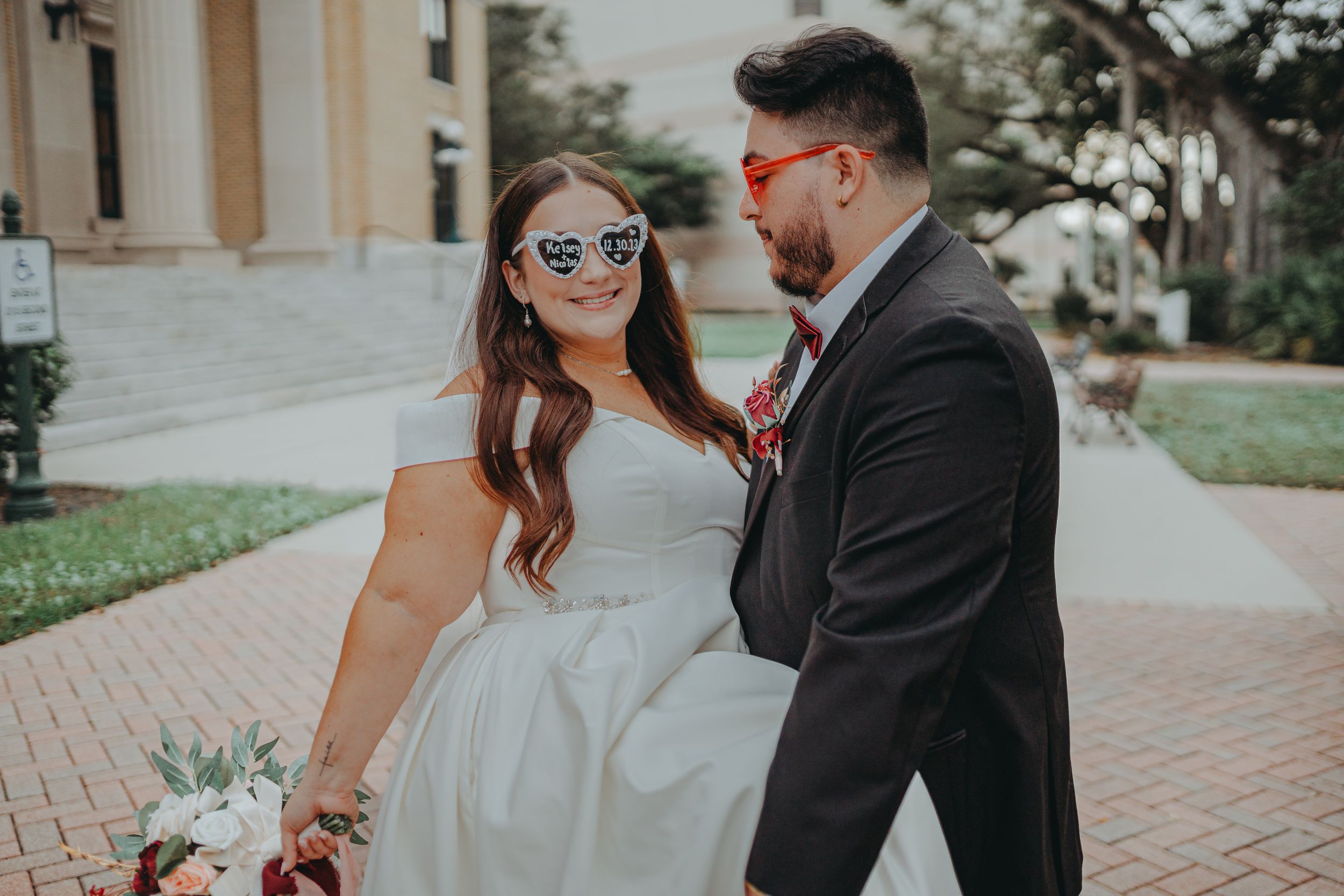Wedding couple in formal attire wearing matching heart-shaped sunglasses on brick pathway.