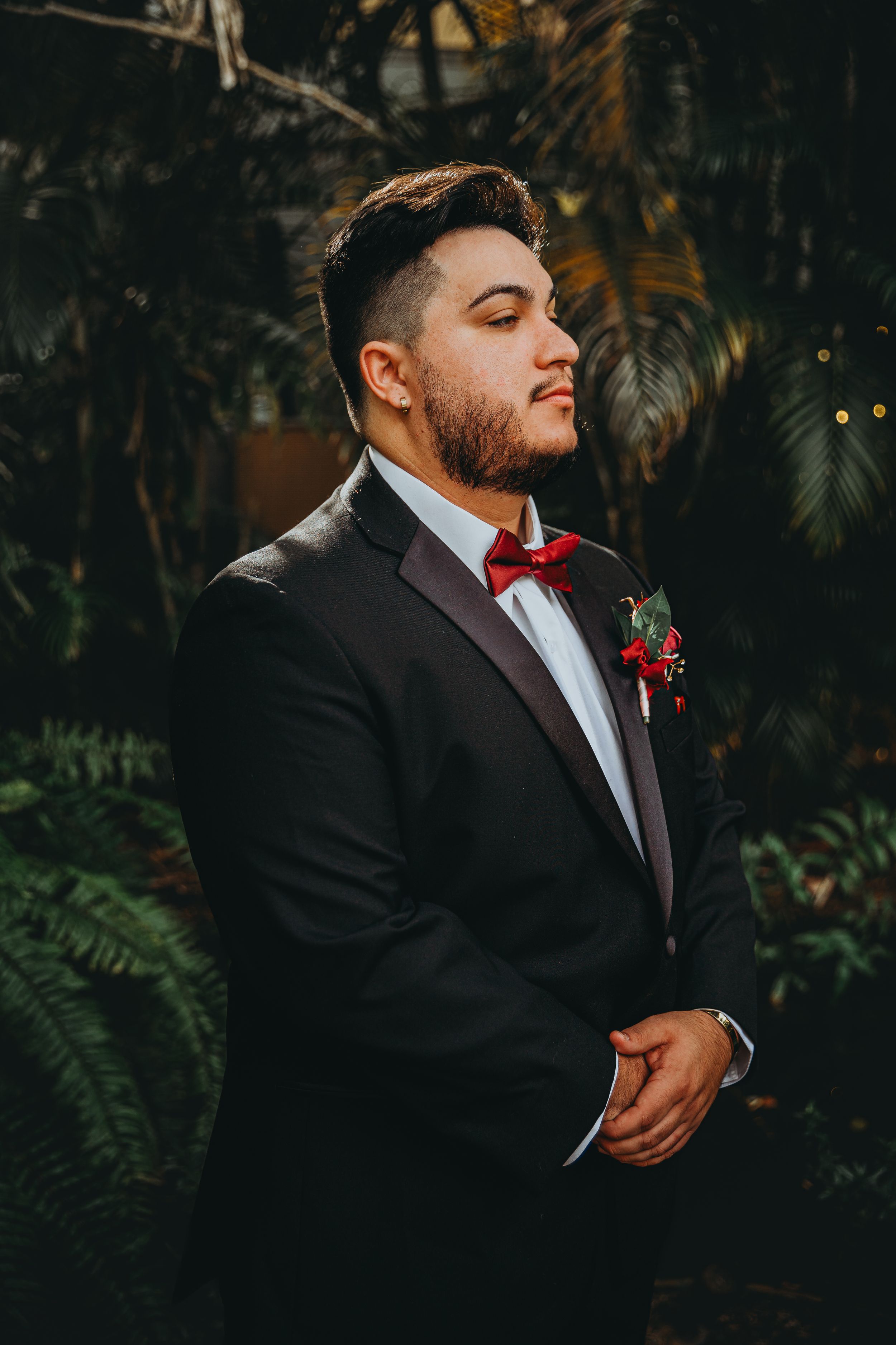 Groom in black tuxedo and red bowtie with boutonniere poses in garden setting.