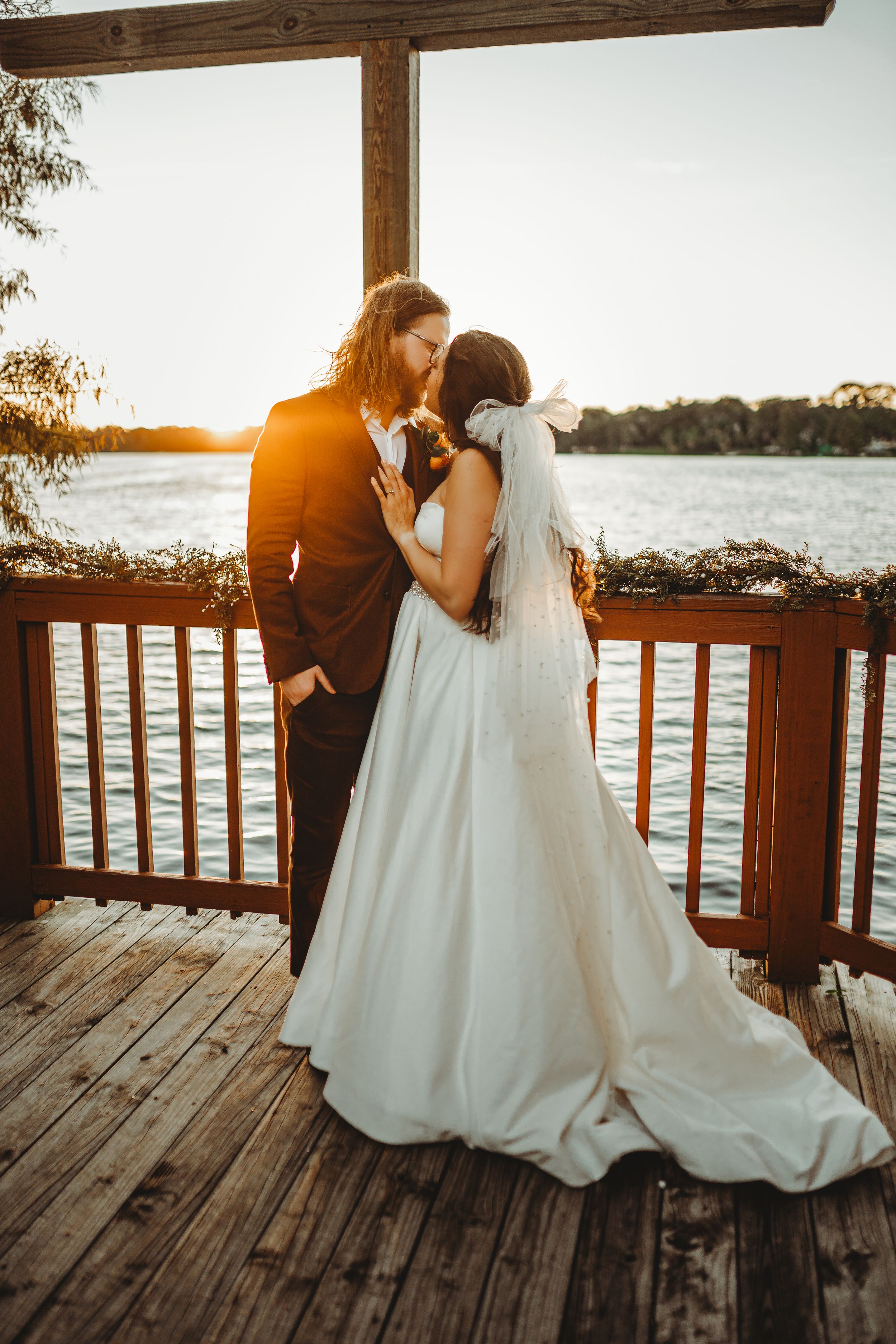 Romantic sunset wedding portrait of couple embracing on wooden deck overlooking water.