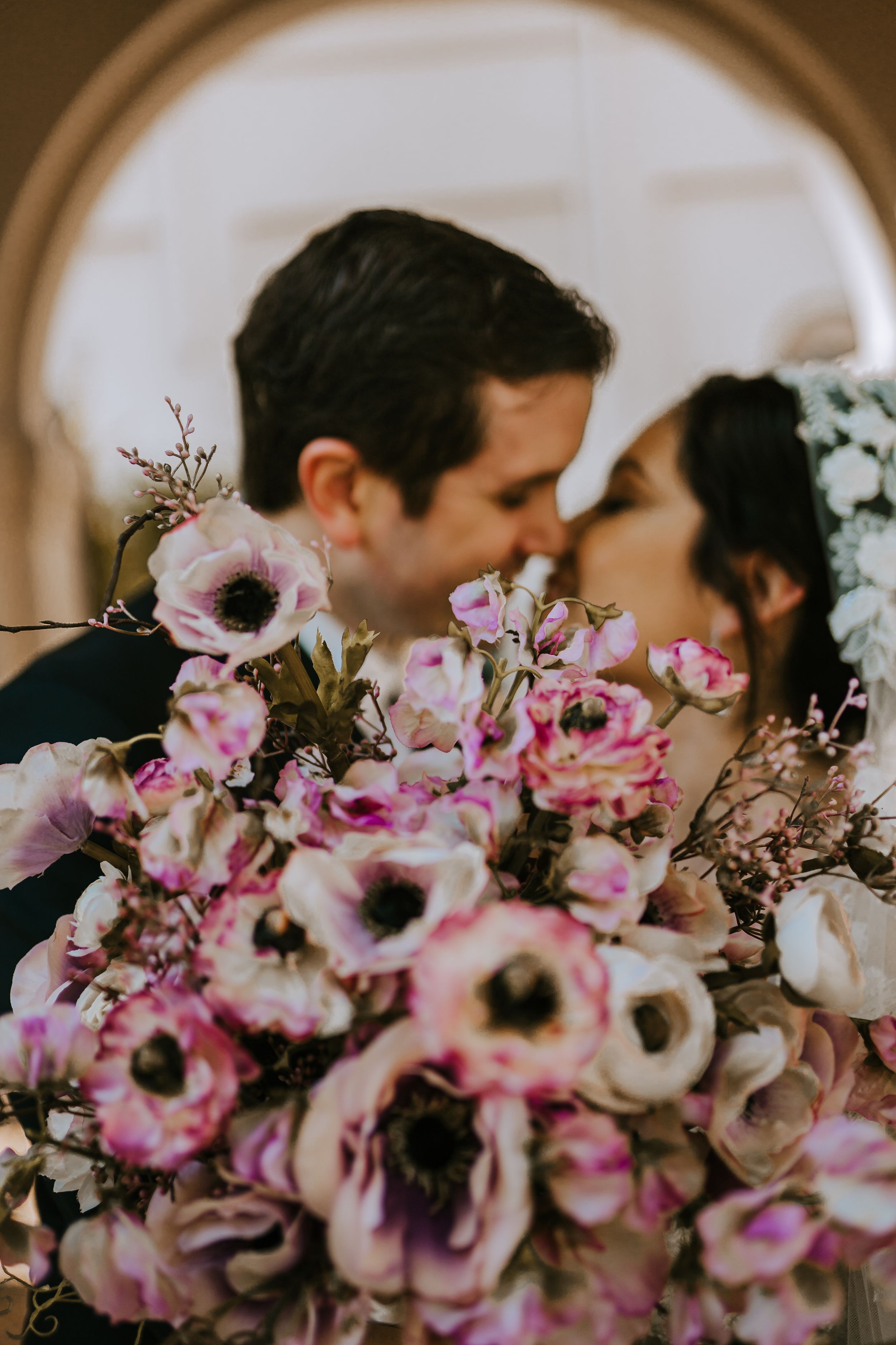 Wedding couple poses behind pink floral bouquet under arched doorway.