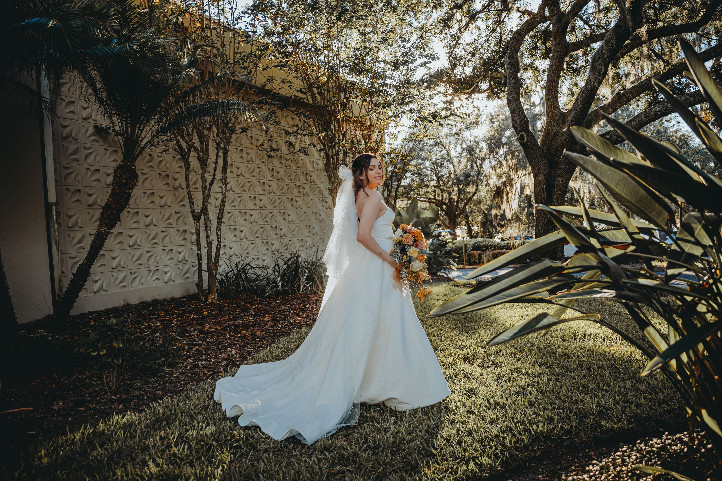 Bride in flowing white gown poses in garden setting with natural sunlight.