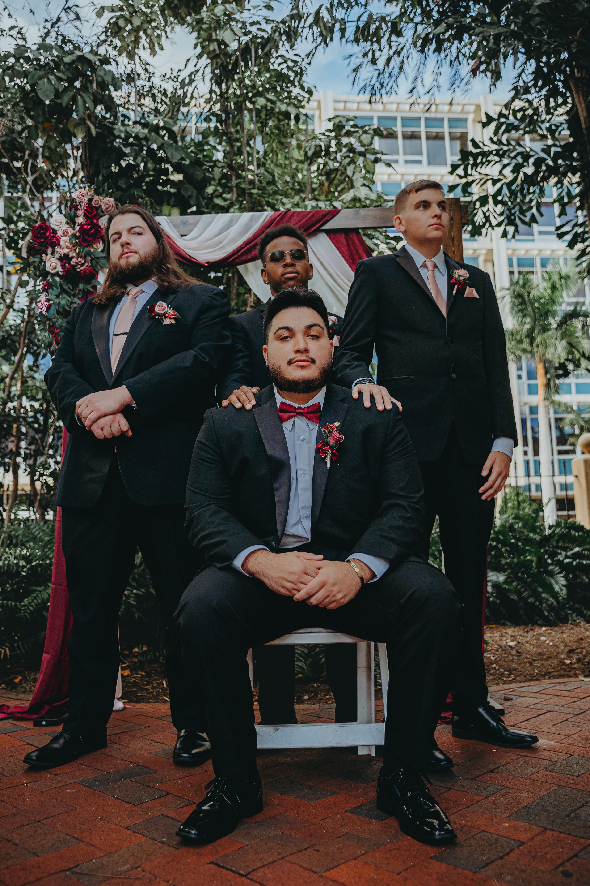 Wedding party in black suits and red bowties pose together outdoors in urban setting.