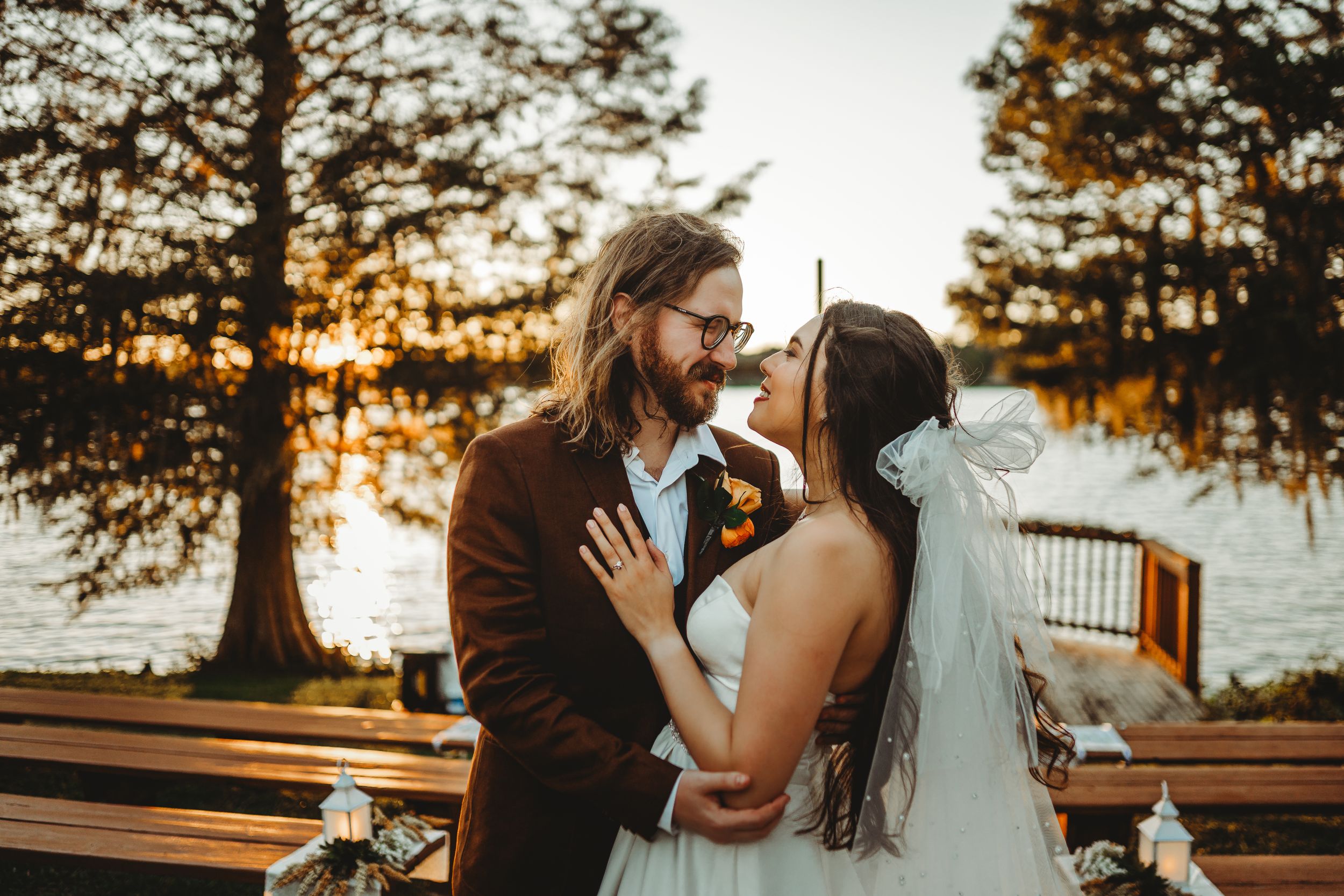 Wedding couple shares intimate moment on wooden deck by lake during golden sunset.