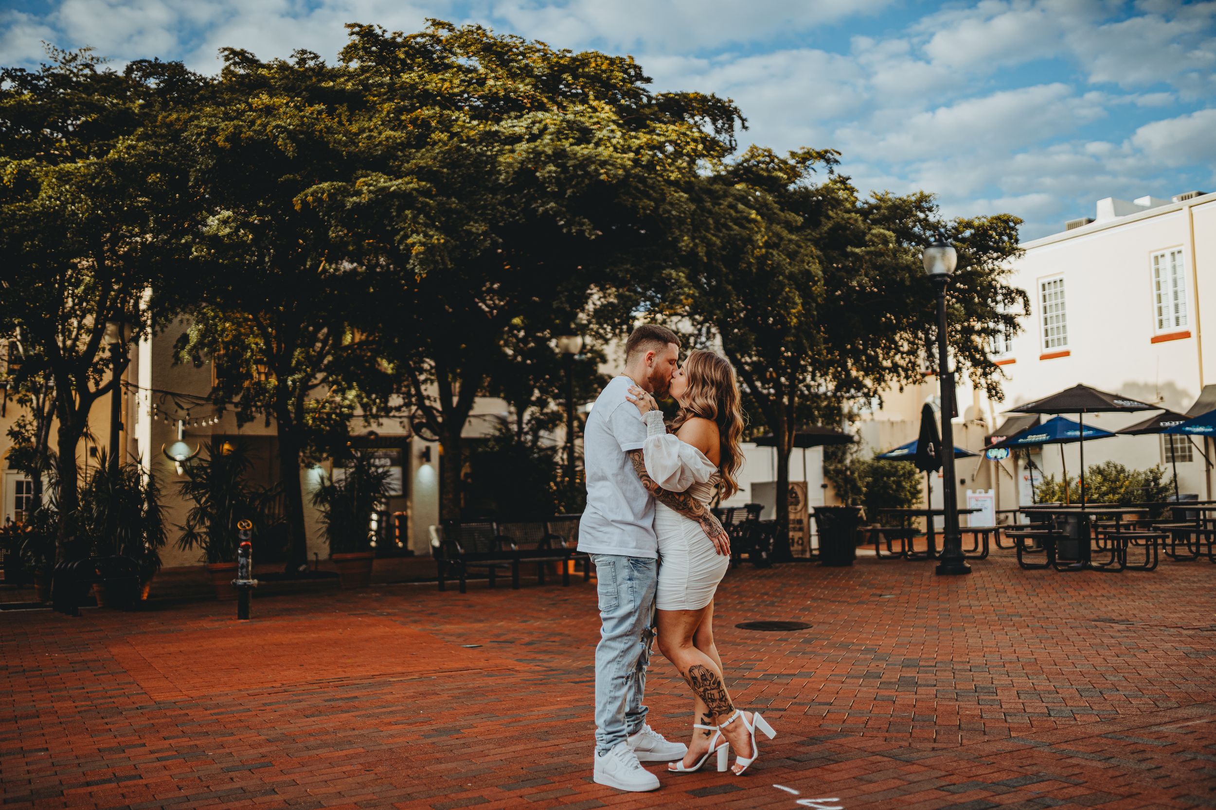 A couple embraces on red brick pavement with lush trees and buildings in the background.