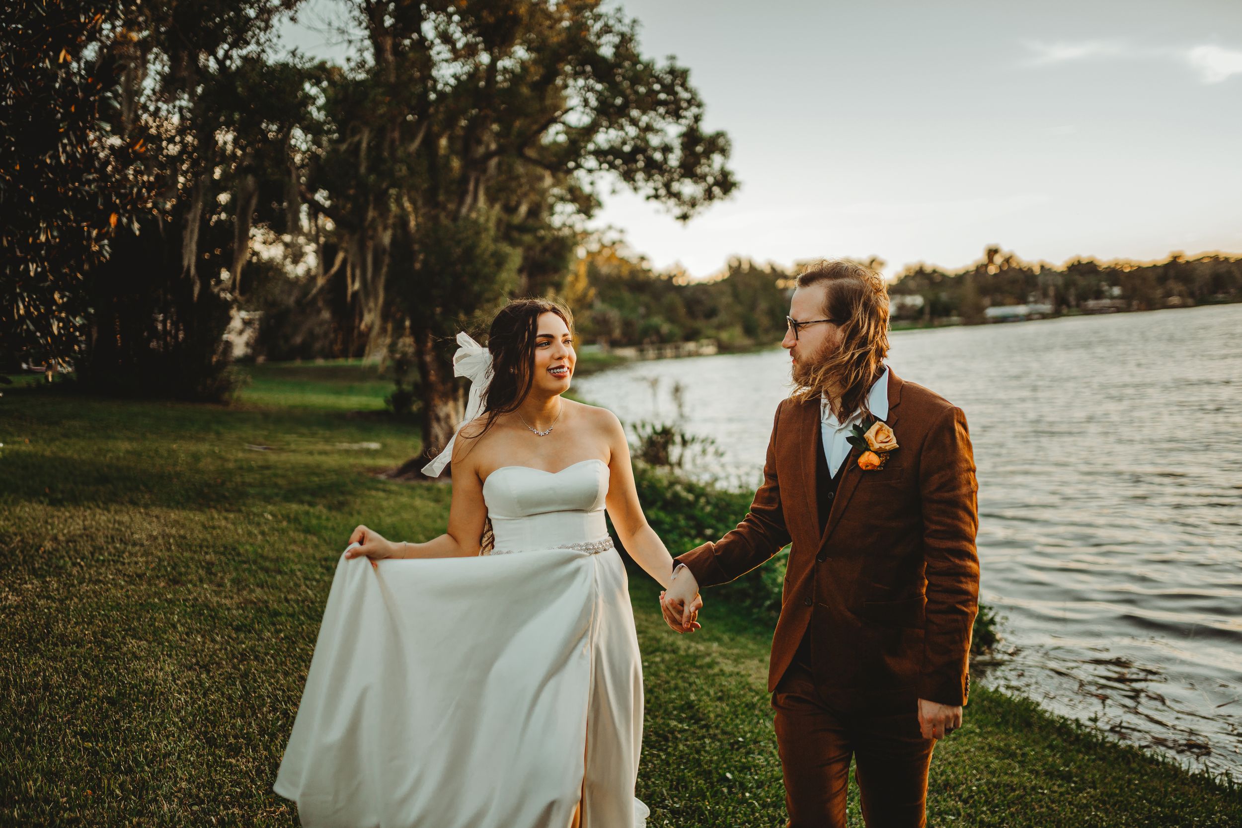 Bride in flowing white dress walks with partner along lakeside path at sunset.