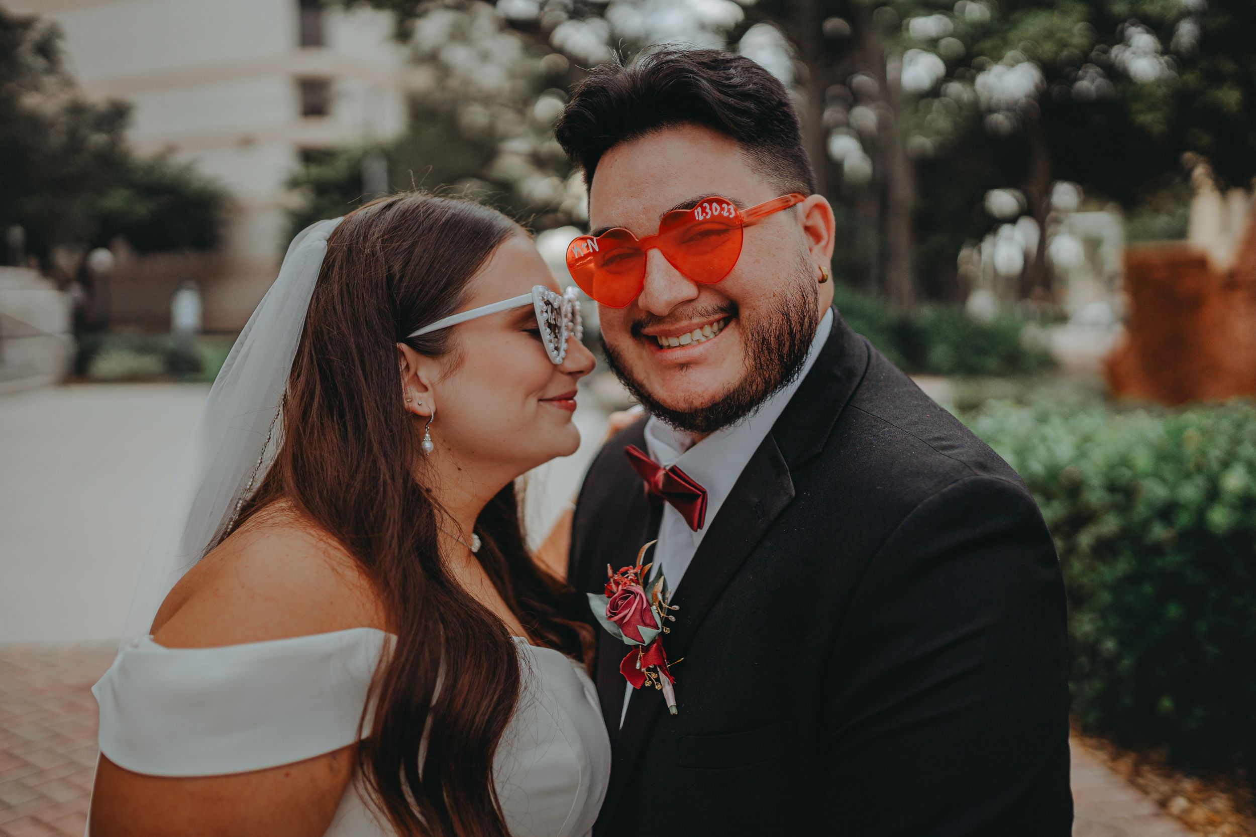 Newlyweds share a laugh while wearing matching heart-shaped sunglasses outdoors.