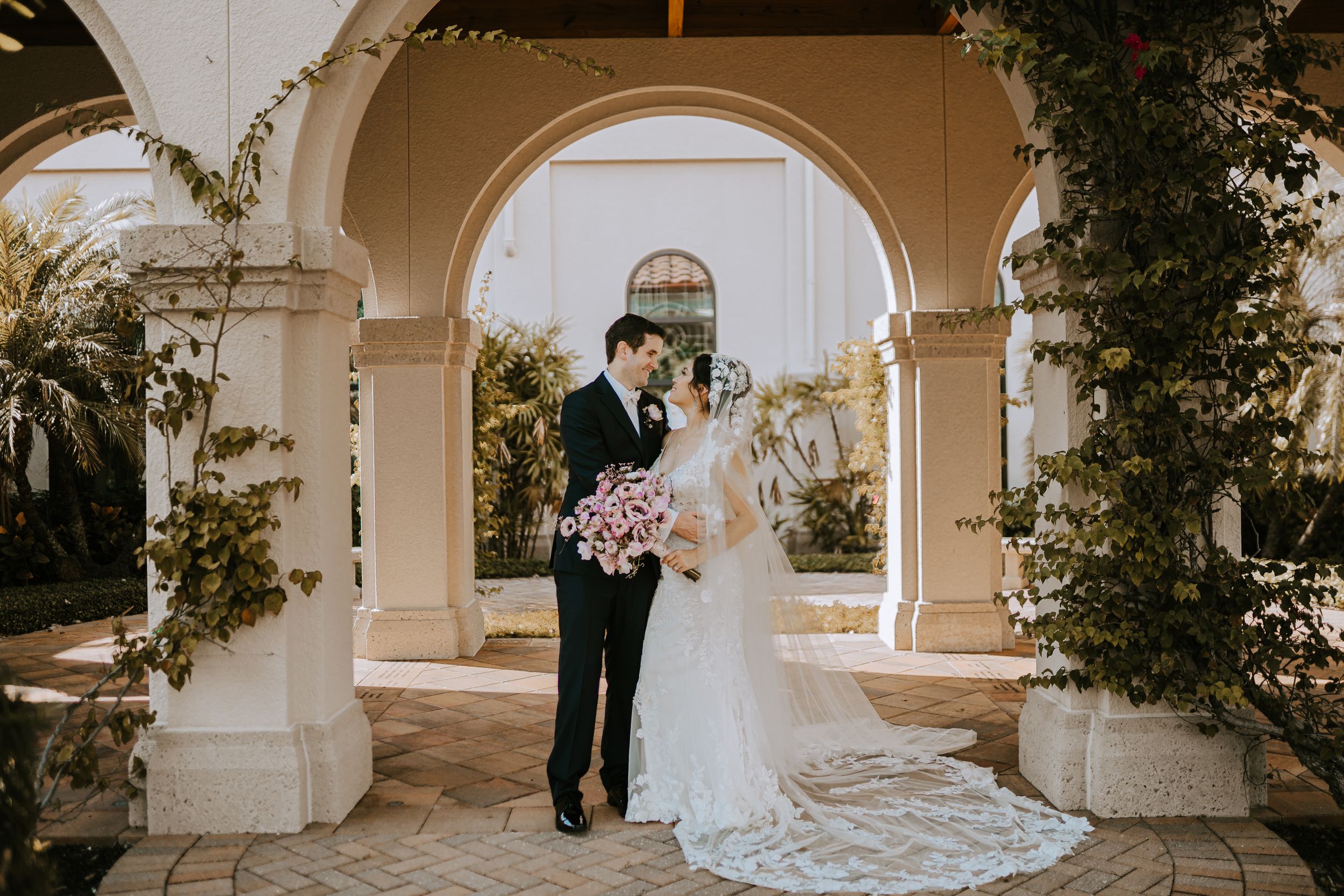 Wedding couple stands under elegant stone archway with classic architecture.