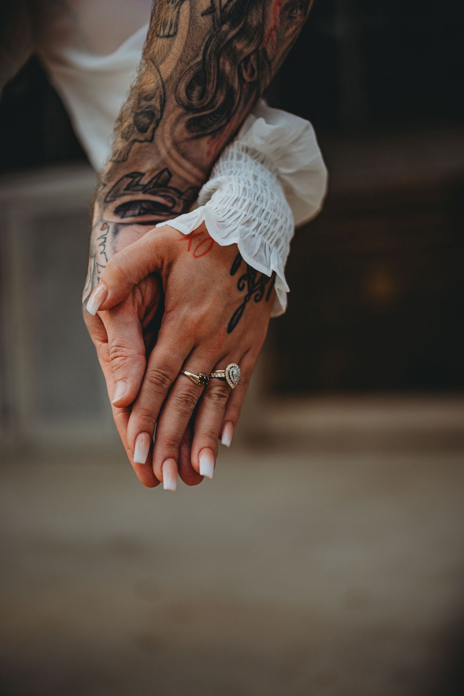 Tattooed hands with engagement ring and white lace sleeve details intertwined against dark background.