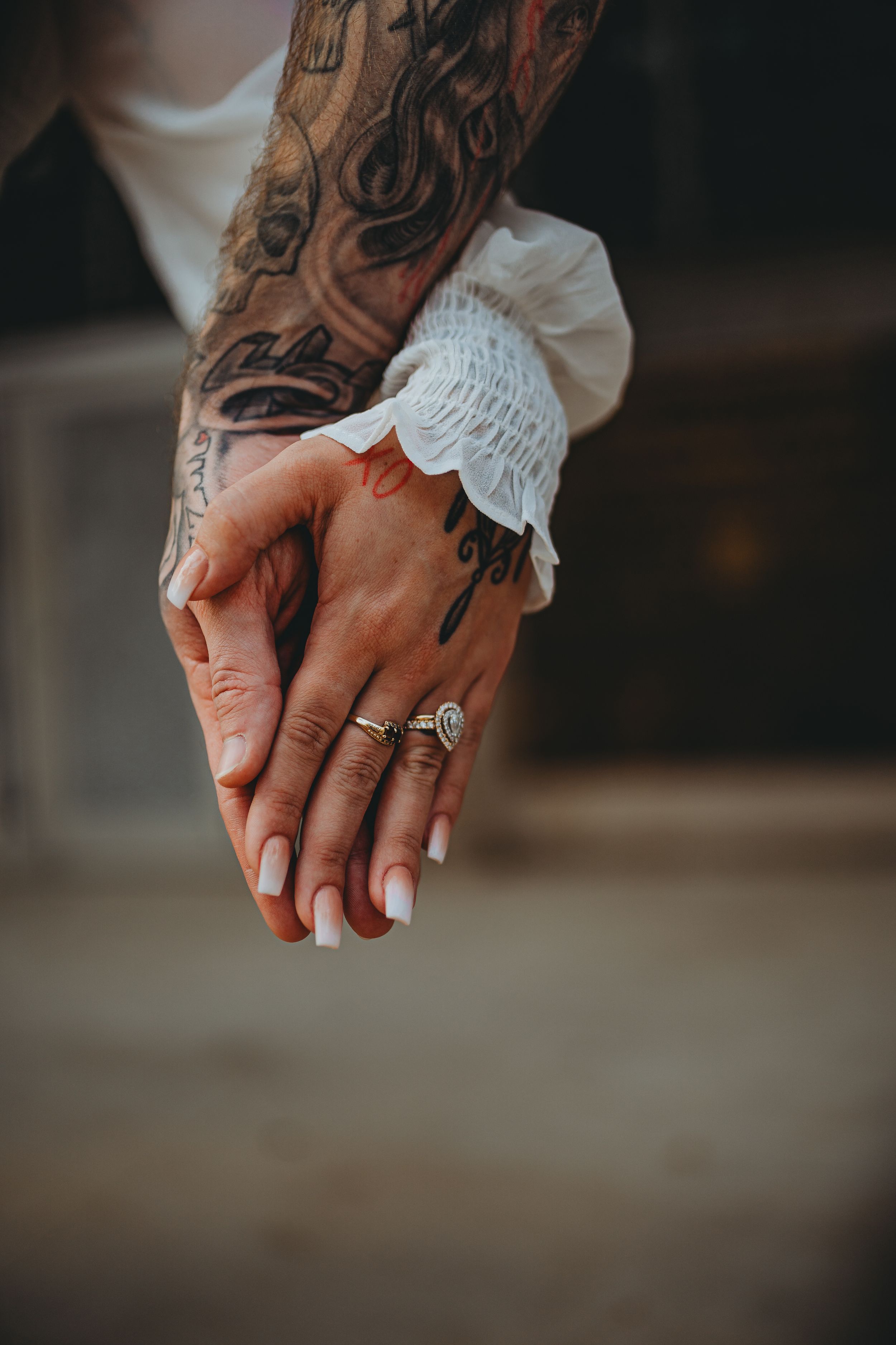 Tattooed hands with engagement ring and white lace sleeve details intertwined against dark background.