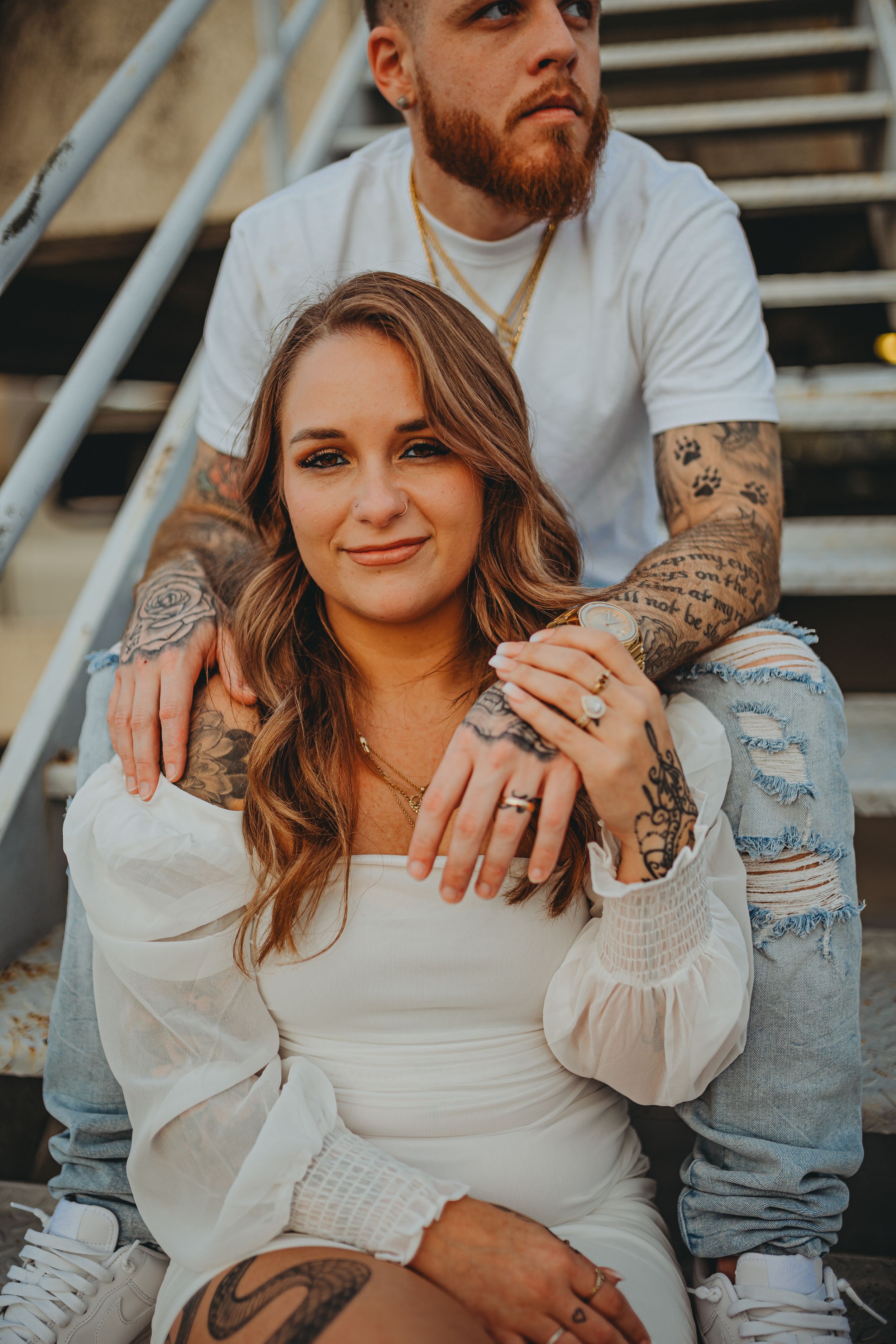 Casual engagement portrait on staircase showing couple's tattoos and ring.