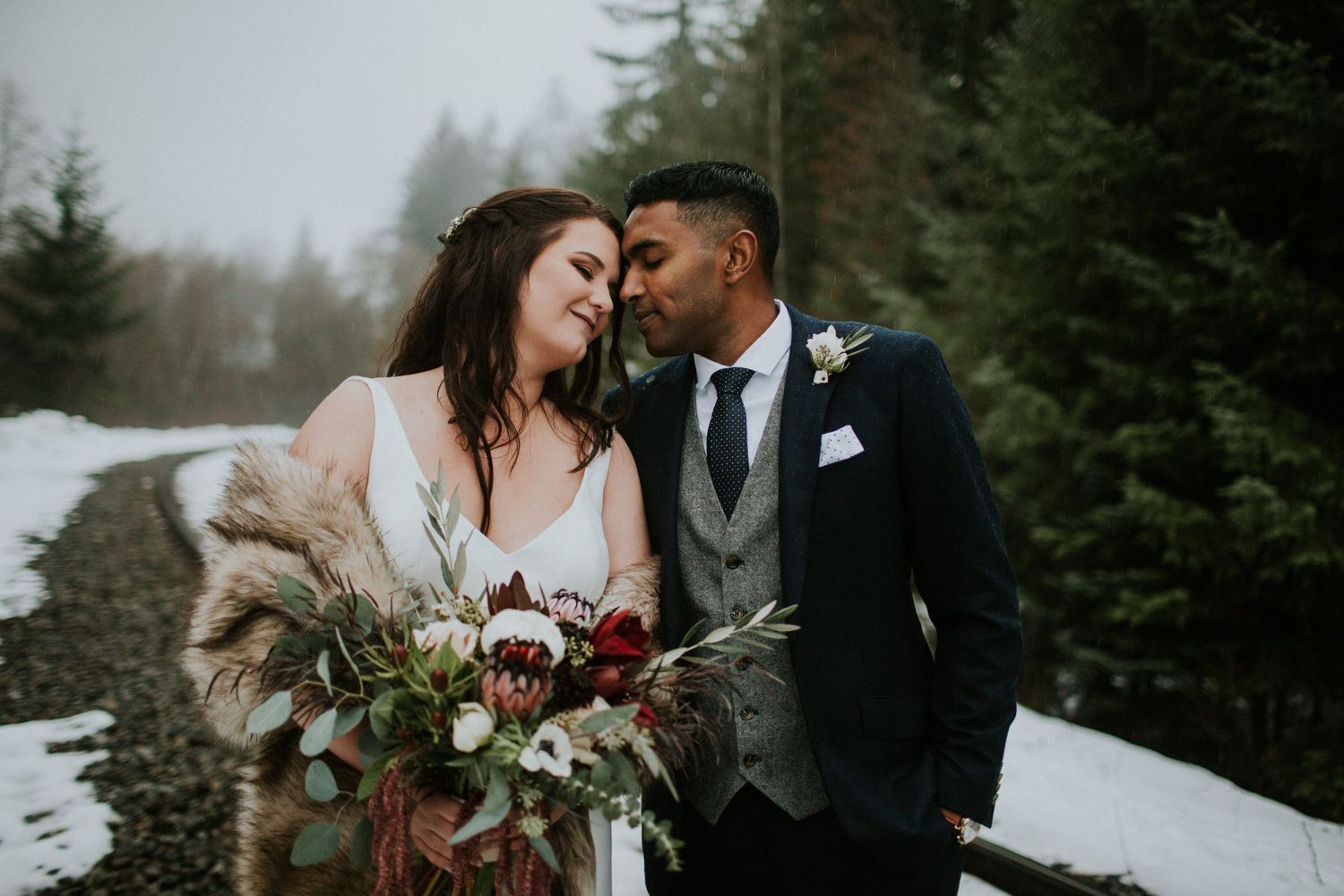 A romantic winter wedding portrait with snow-covered evergreens in the background.