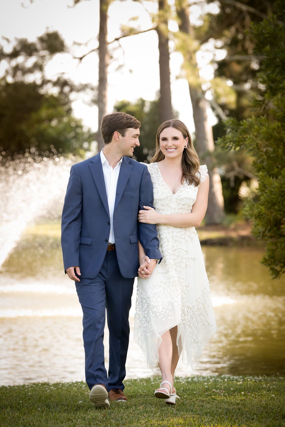 Couple in wedding attire walks together on grass near a waterfront at golden hour.