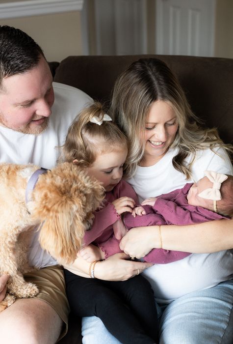 Family cuddles on brown couch with goldendoodle dog while meeting their newborn baby.
