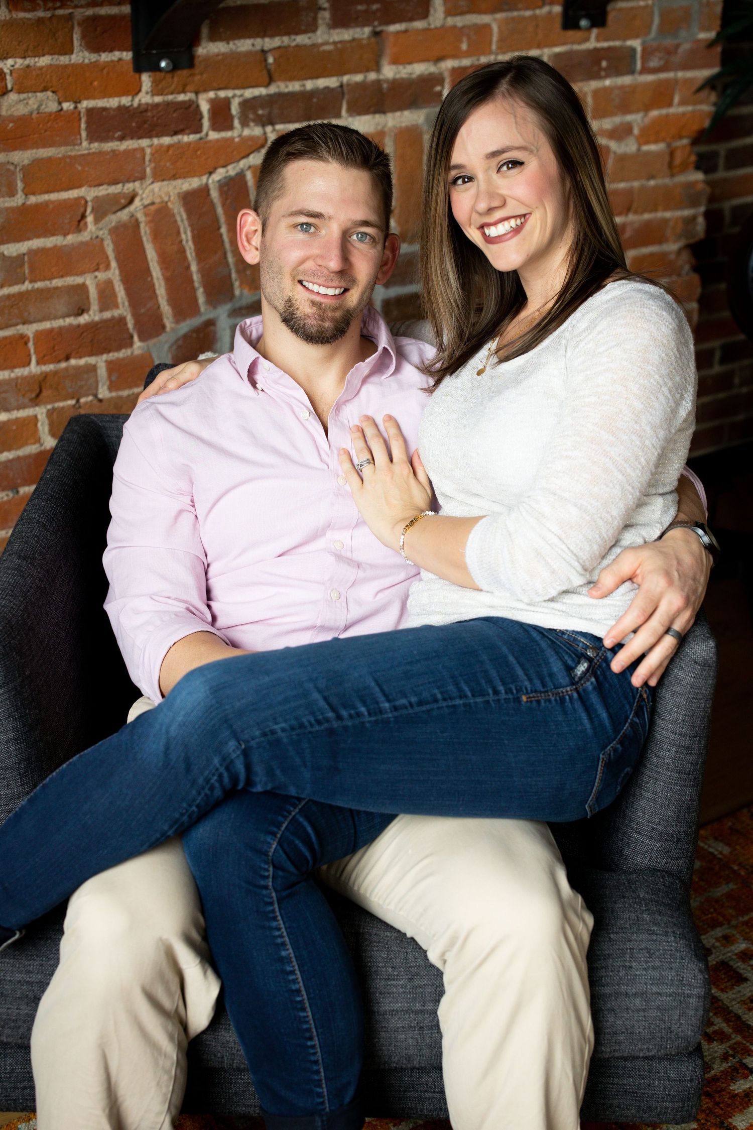 Couple relaxes on grey couch against brick wall during casual photo session.