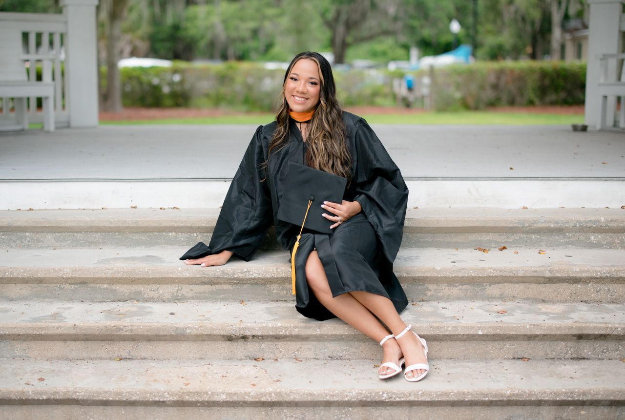 A graduation photo of someone in a black cap and gown sitting on concrete steps outdoors.