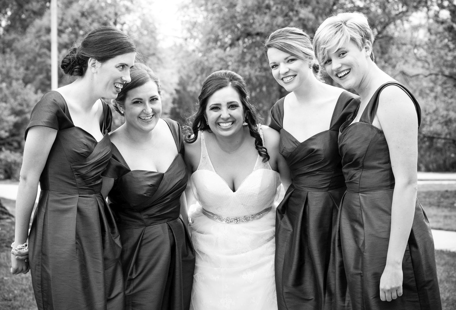 A group of bridesmaids in matching black dresses pose with the bride at an outdoor wedding.