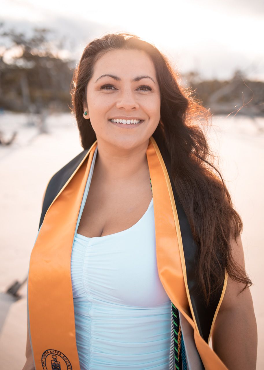 A graduation portrait of someone wearing an orange stole against a bright sunset background.