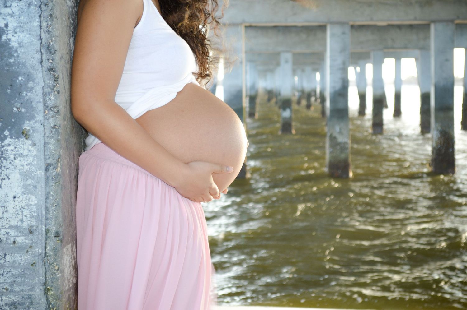 Side view of pregnant woman in pink skirt standing near pier supports over water.