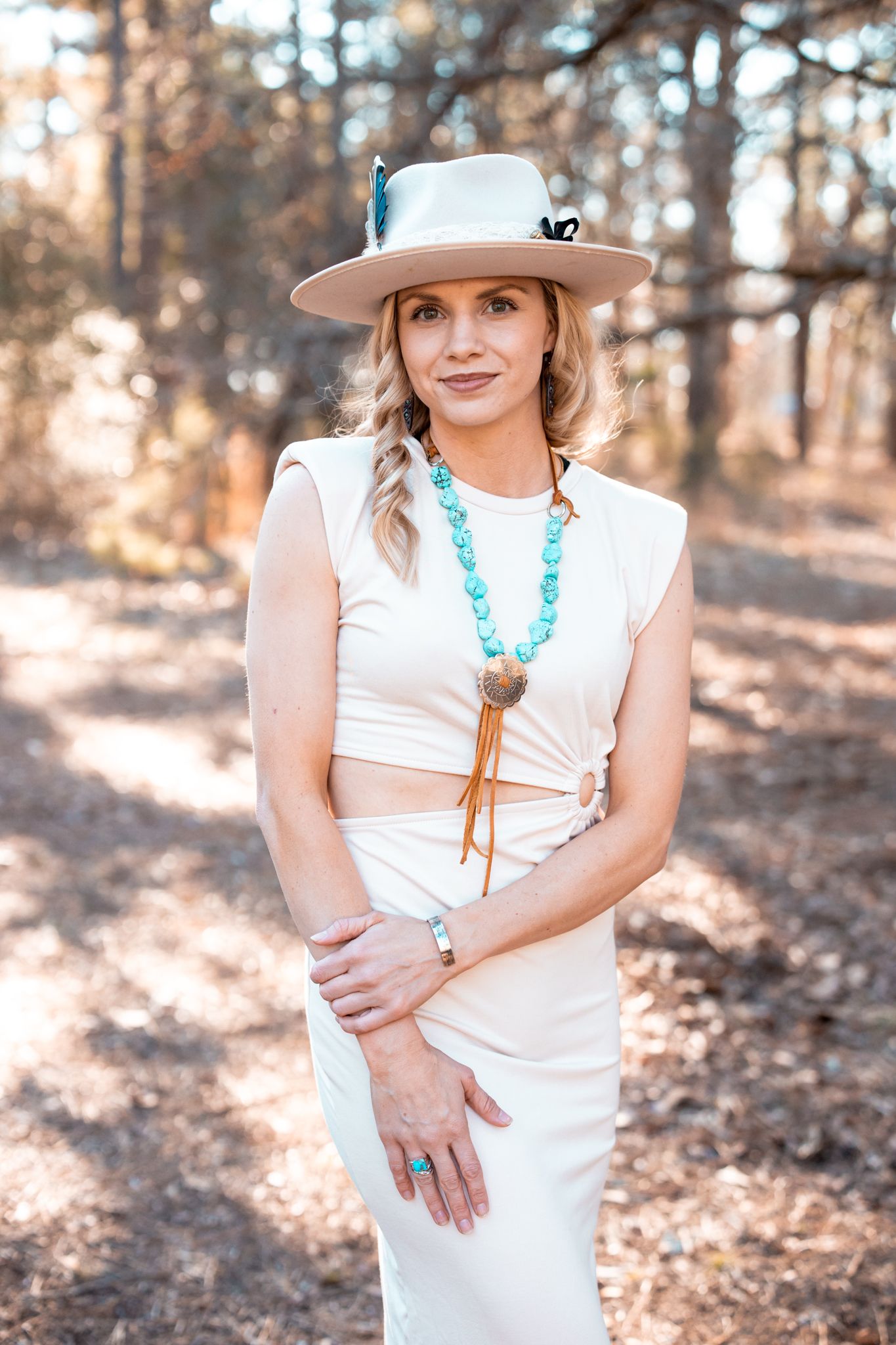 Woman in white dress and tan hat poses in autumn forest setting with turquoise jewelry accents.