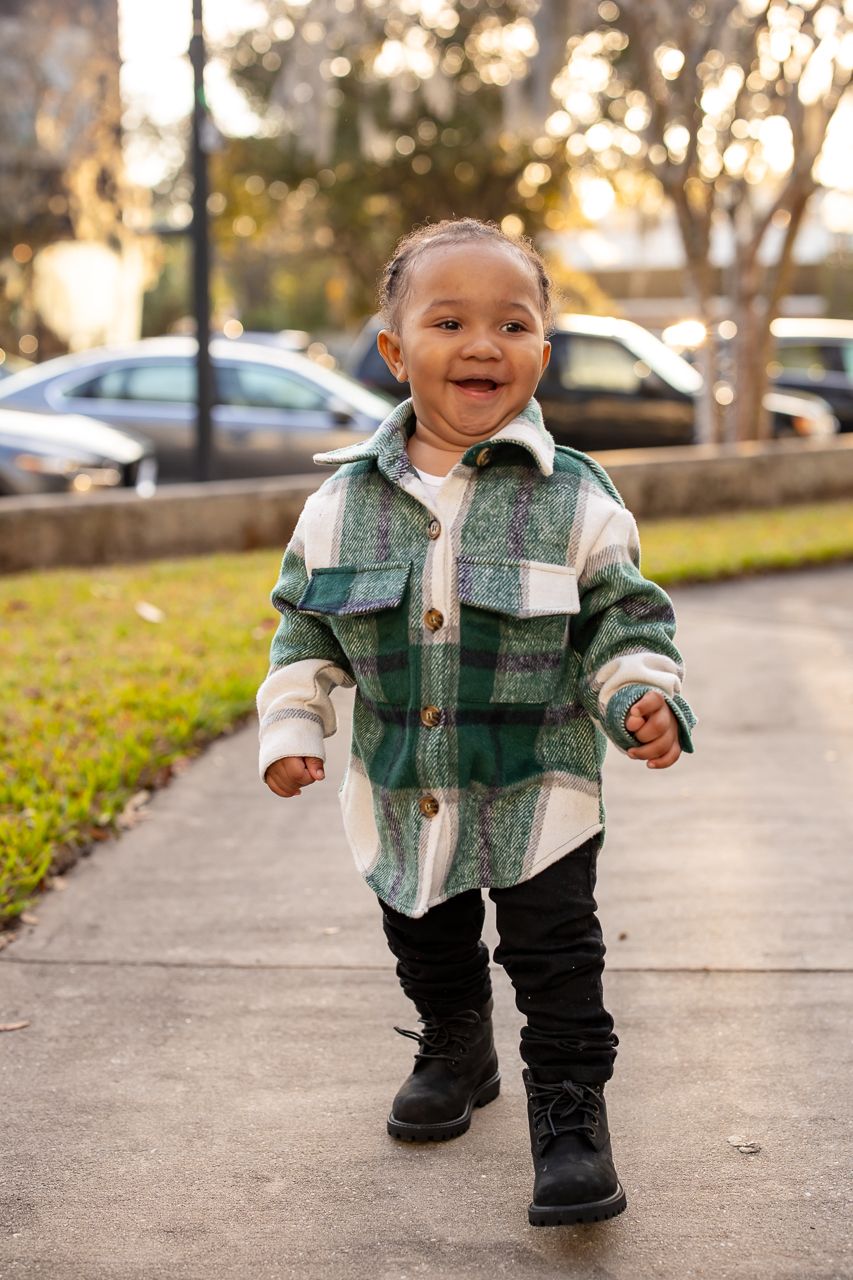 Toddler walking outdoors wearing a green plaid jacket and dark pants.