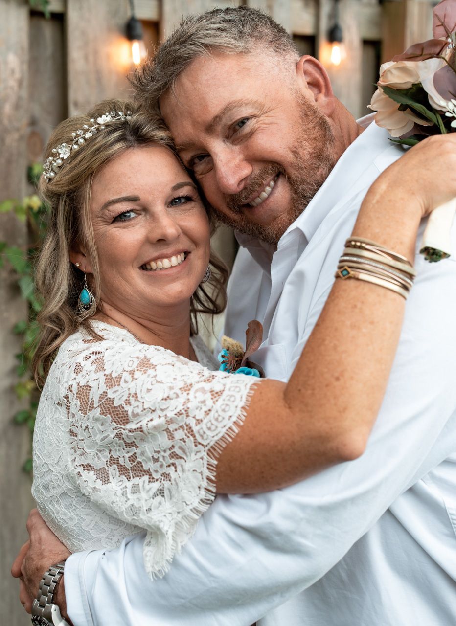 A wedding couple in white embracing and smiling with decorative lights in the background.