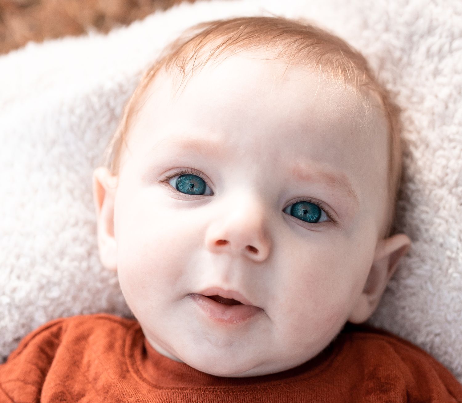 A baby with bright blue eyes wearing an orange outfit smiles while lying on a white blanket.