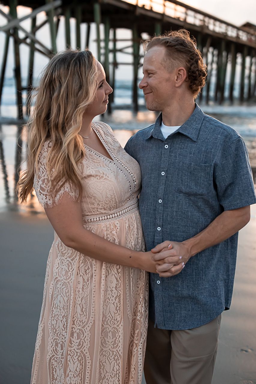Couple poses together near pier at sunset wearing blue and blush tones.