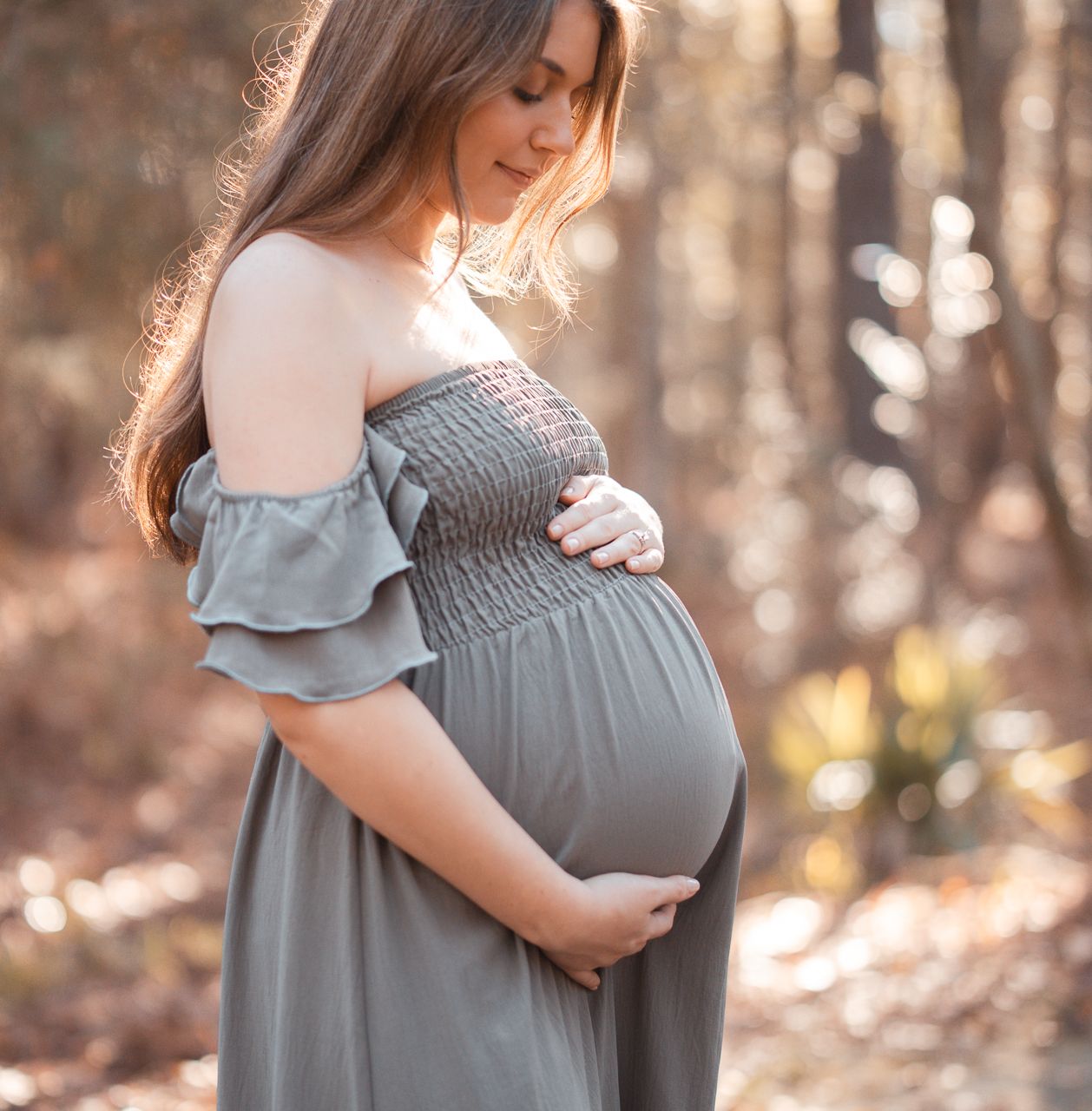 Maternity photos in a grey dress with soft natural lighting in a wooded setting.