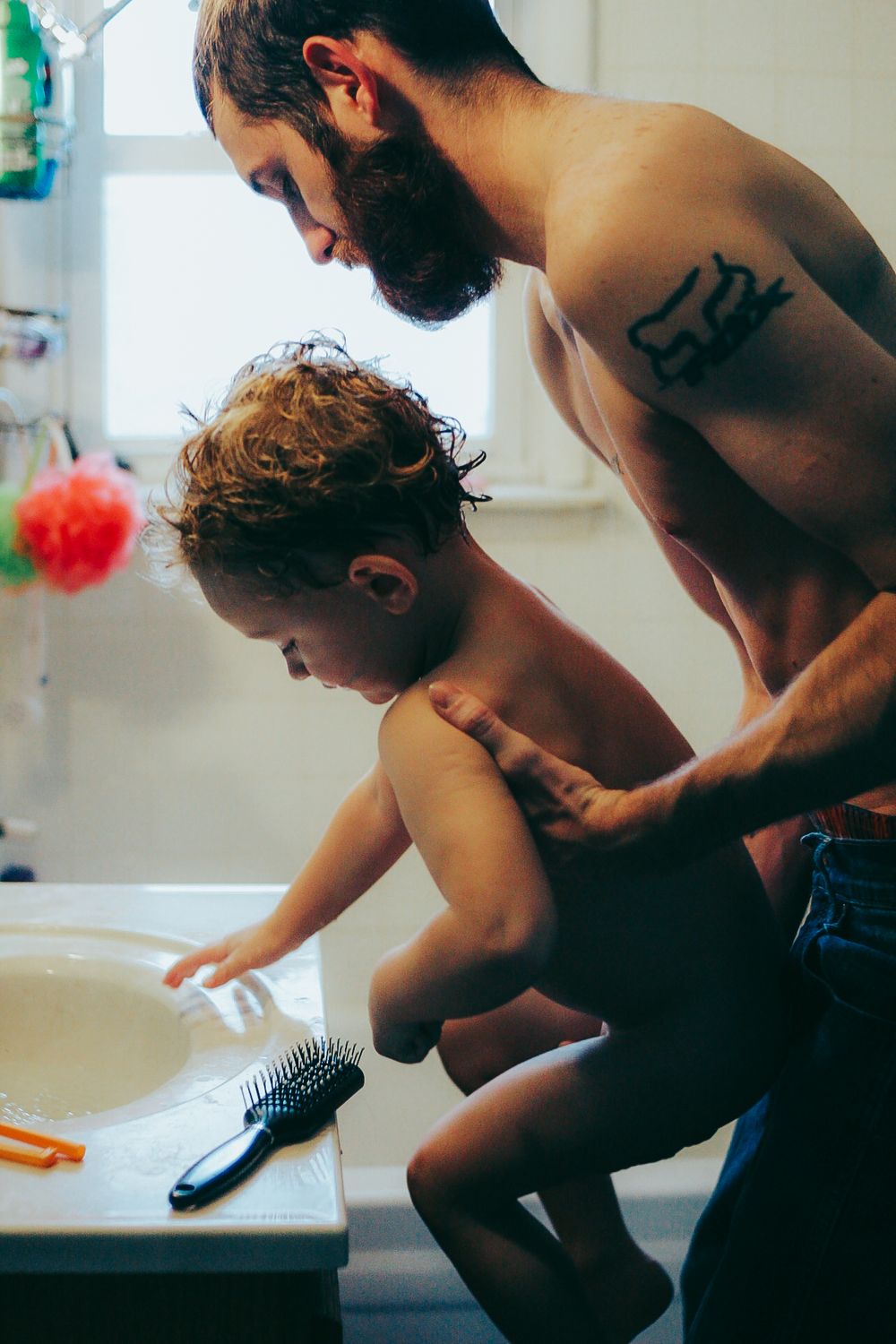 A bearded man helps a small child lean over a bathroom sink.