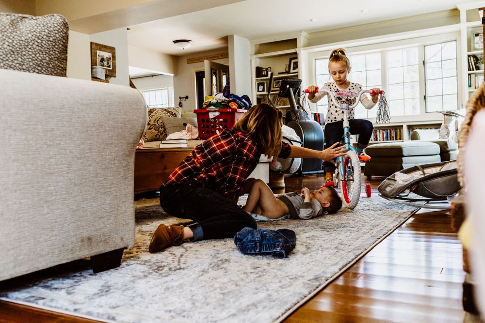 Family gathers around a small bicycle on a light gray rug in modern living room.