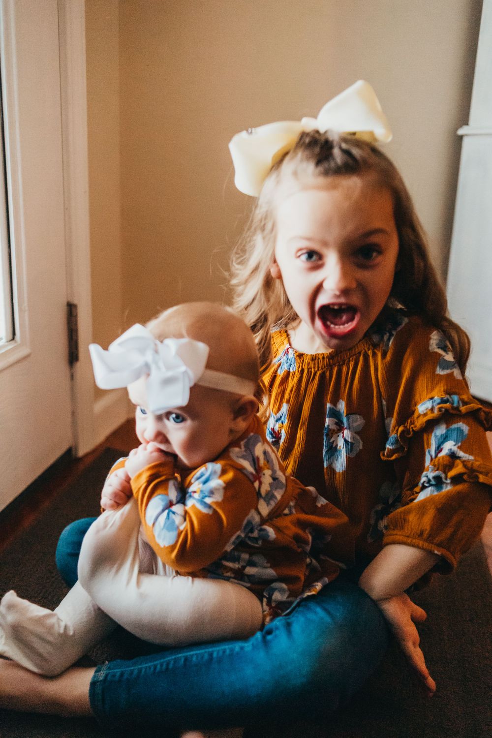Two children in matching mustard floral outfits and white bows sitting together indoors.