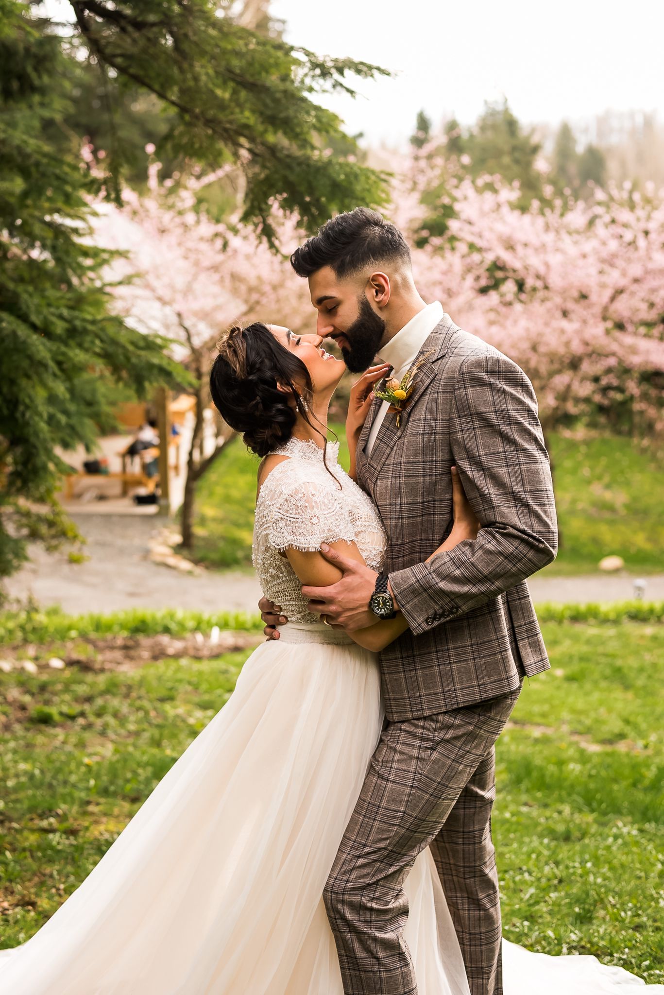 In front of the cherry blossom trees, a couple smiles at each other as they celebrate their elopement at a small greenhouse.