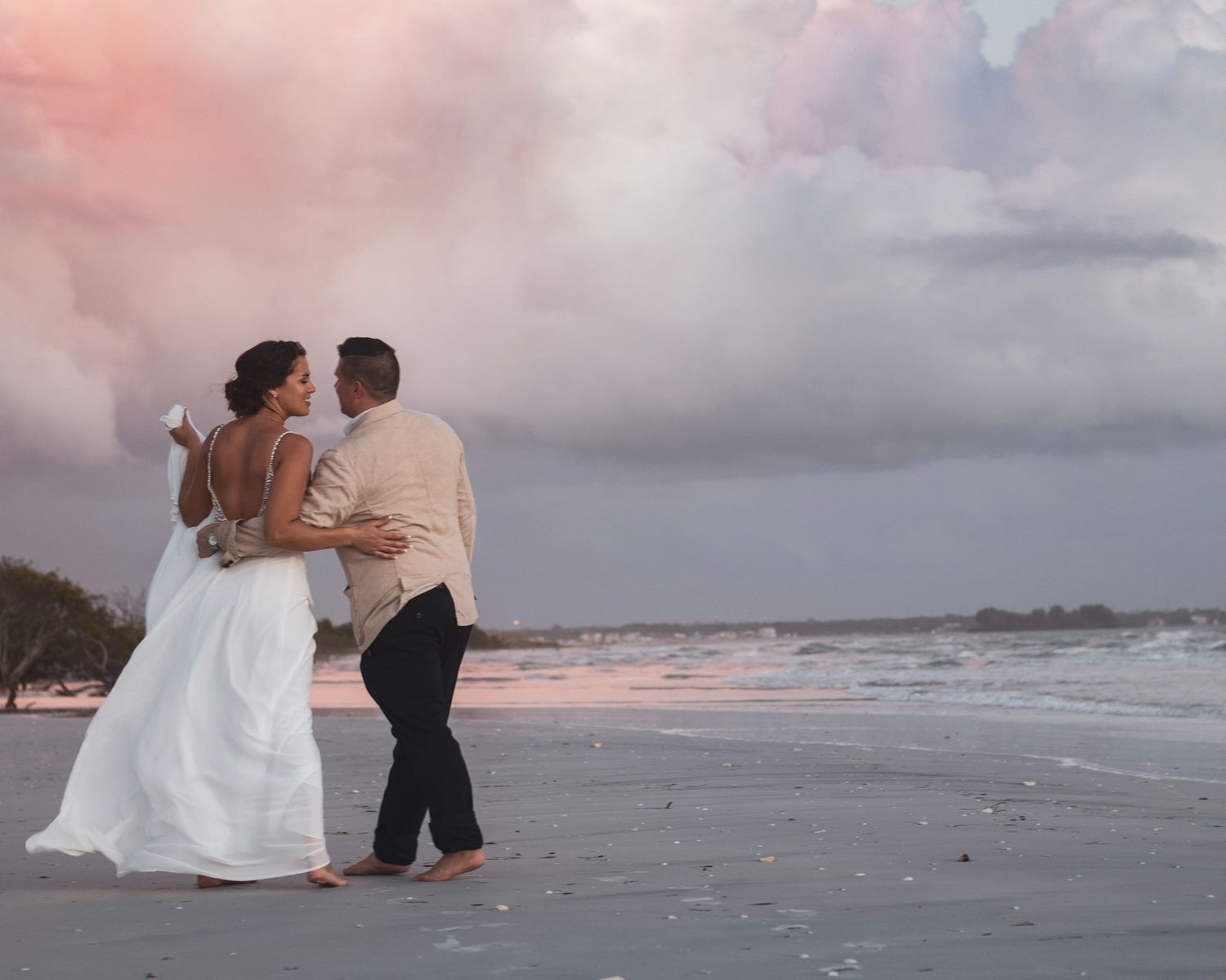 A couple shares an intimate moment on a beach at sunset with pink clouds.