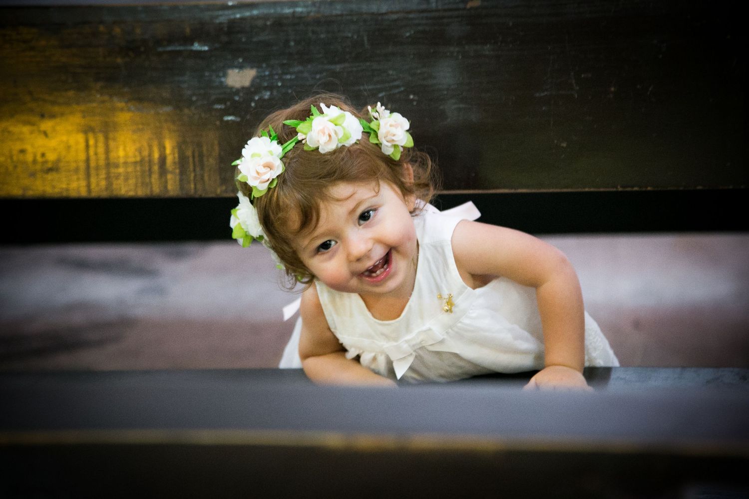 Adorable smiling toddler girl wearing a floral crown leans over a wooden bench.