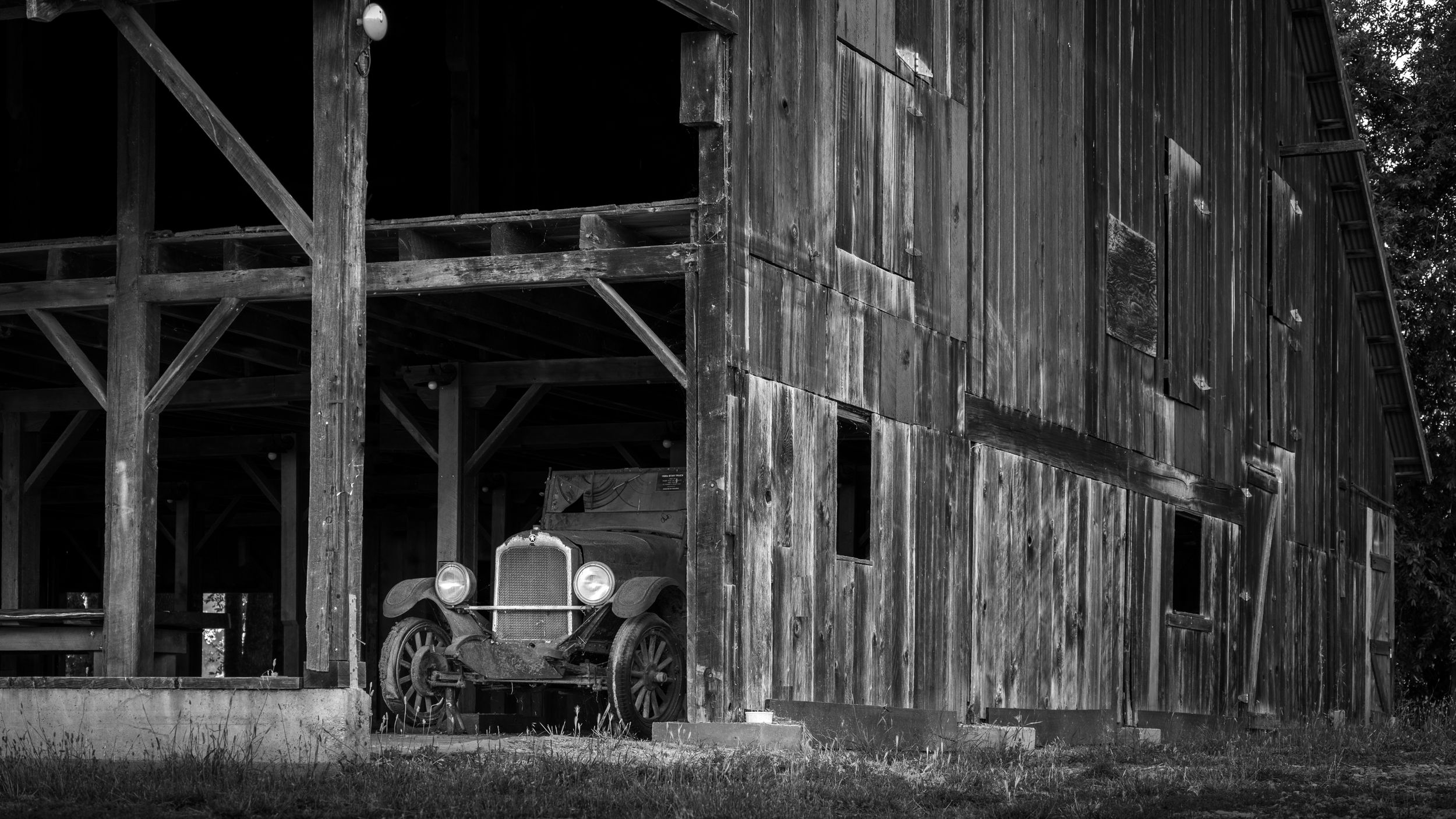 Vintage car parked next to rustic wooden barn in black and white.