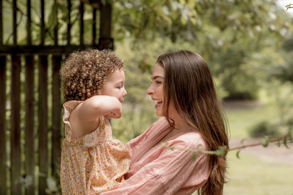 A smiling mother holds her curly-haired toddler face-to-face in a lush green garden.